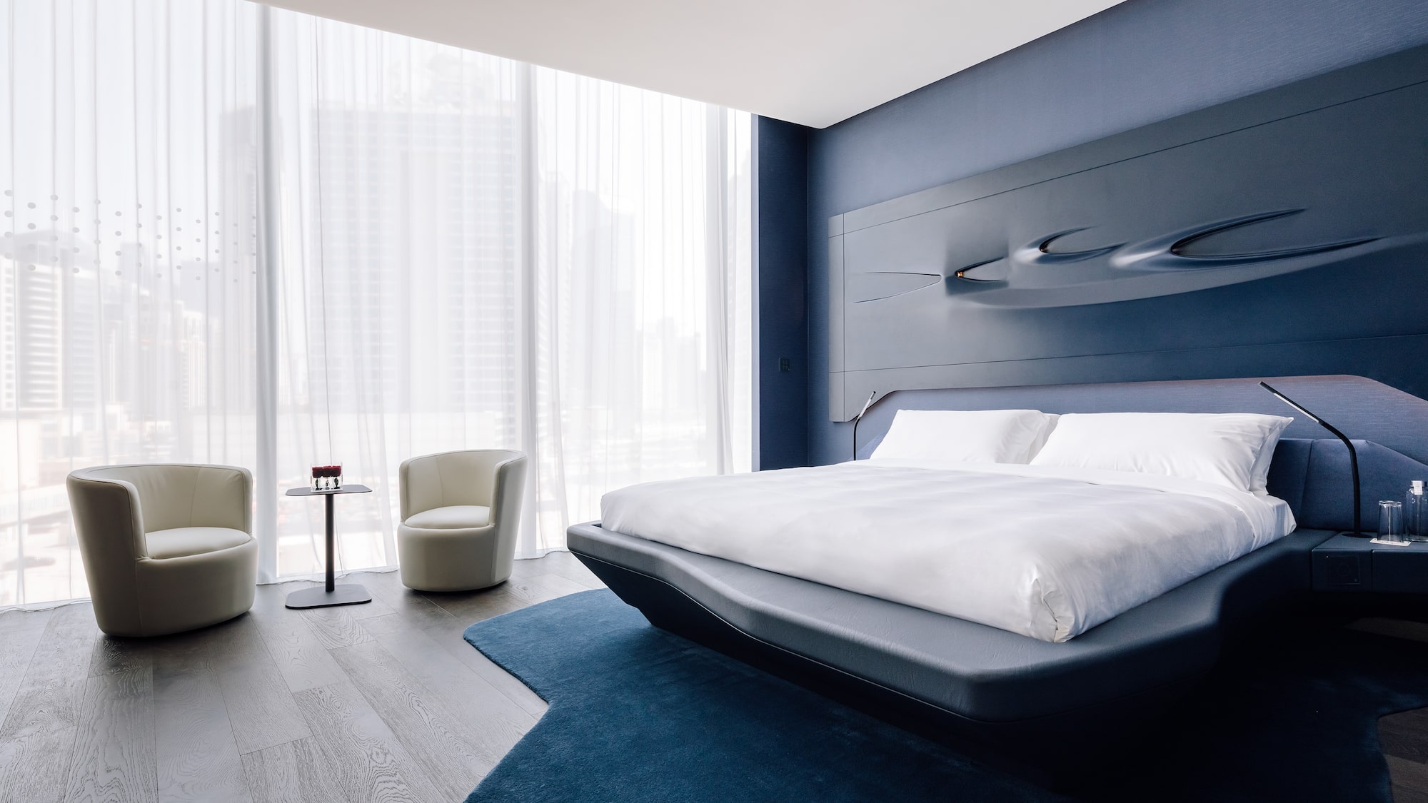 Modern blue bedroom with sculptural headboard, city view, and contemporary seating.