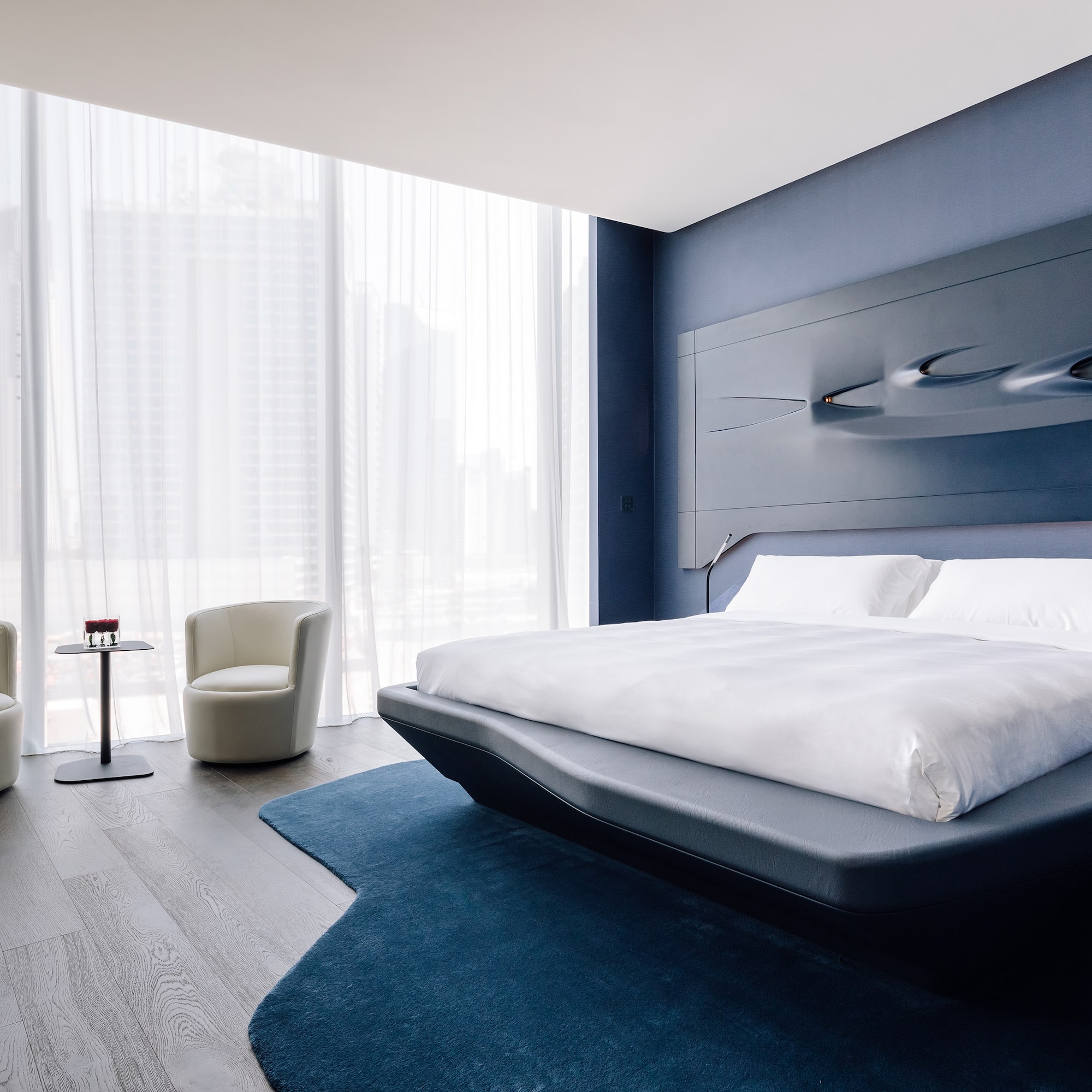 Modern blue bedroom with sculptural headboard, city view, and contemporary seating.
