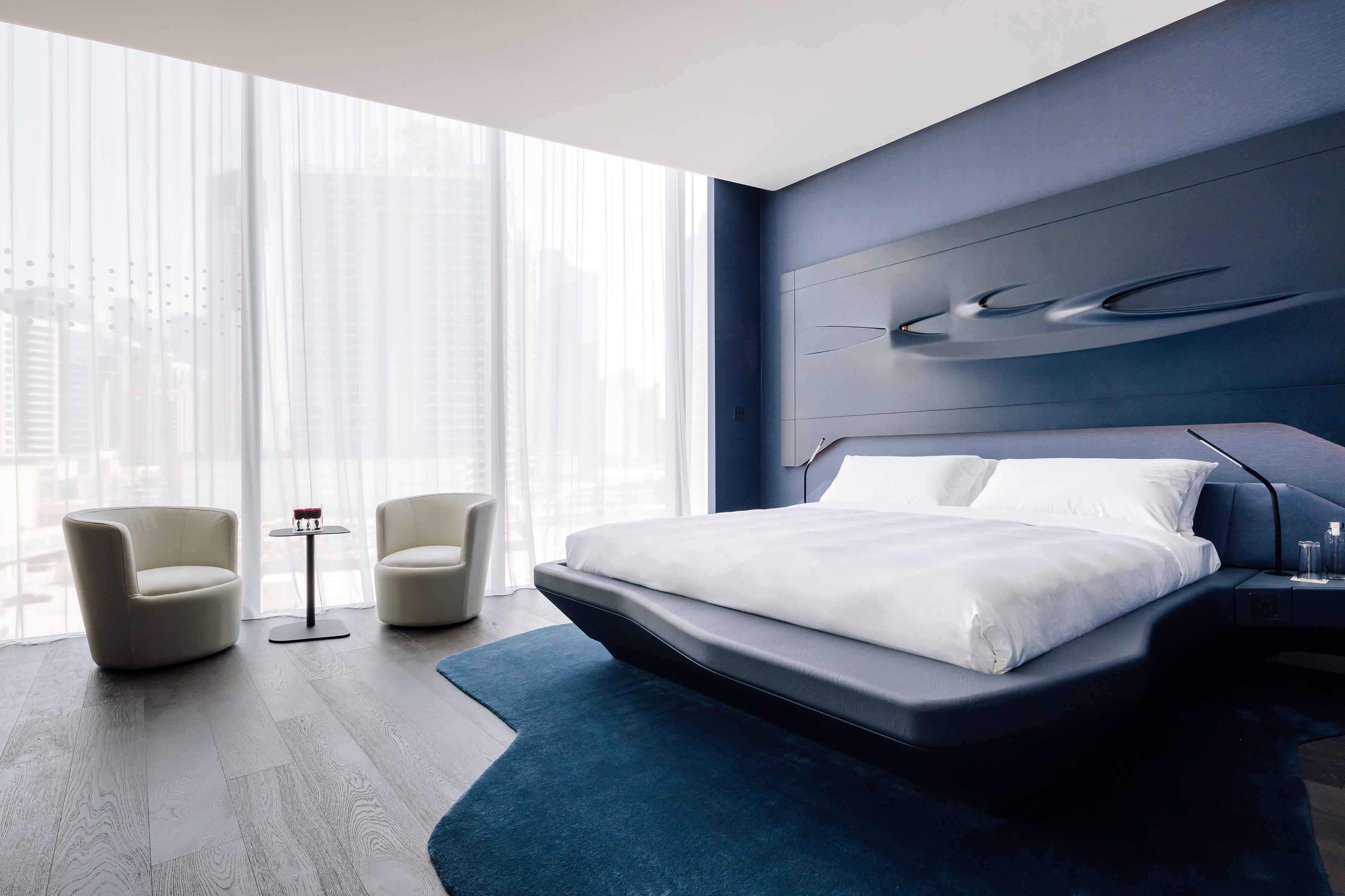 Modern blue bedroom with sculptural headboard, city view, and contemporary seating.