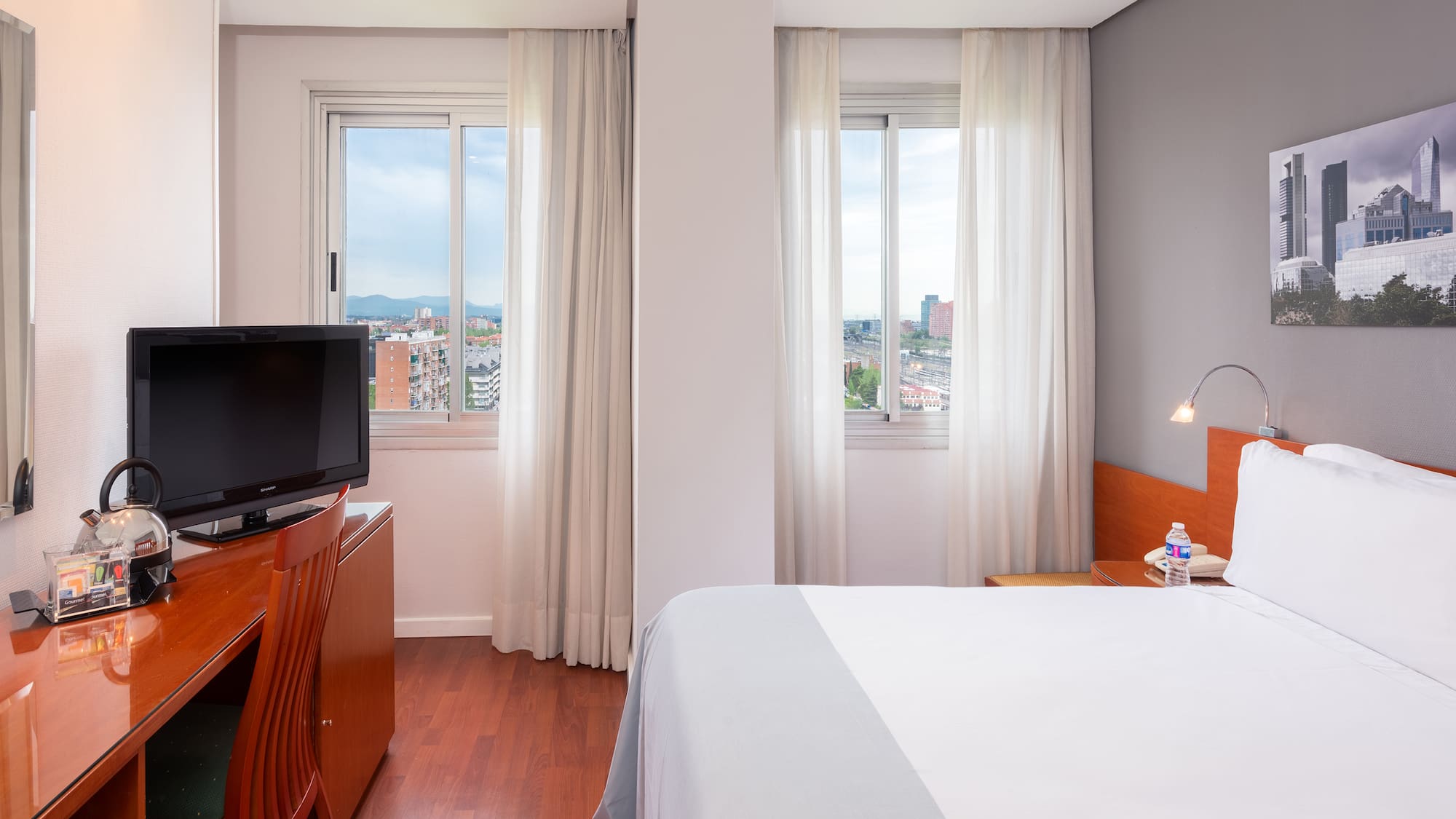 Modern hotel room with city view, comfortable bed, desk, and TV.