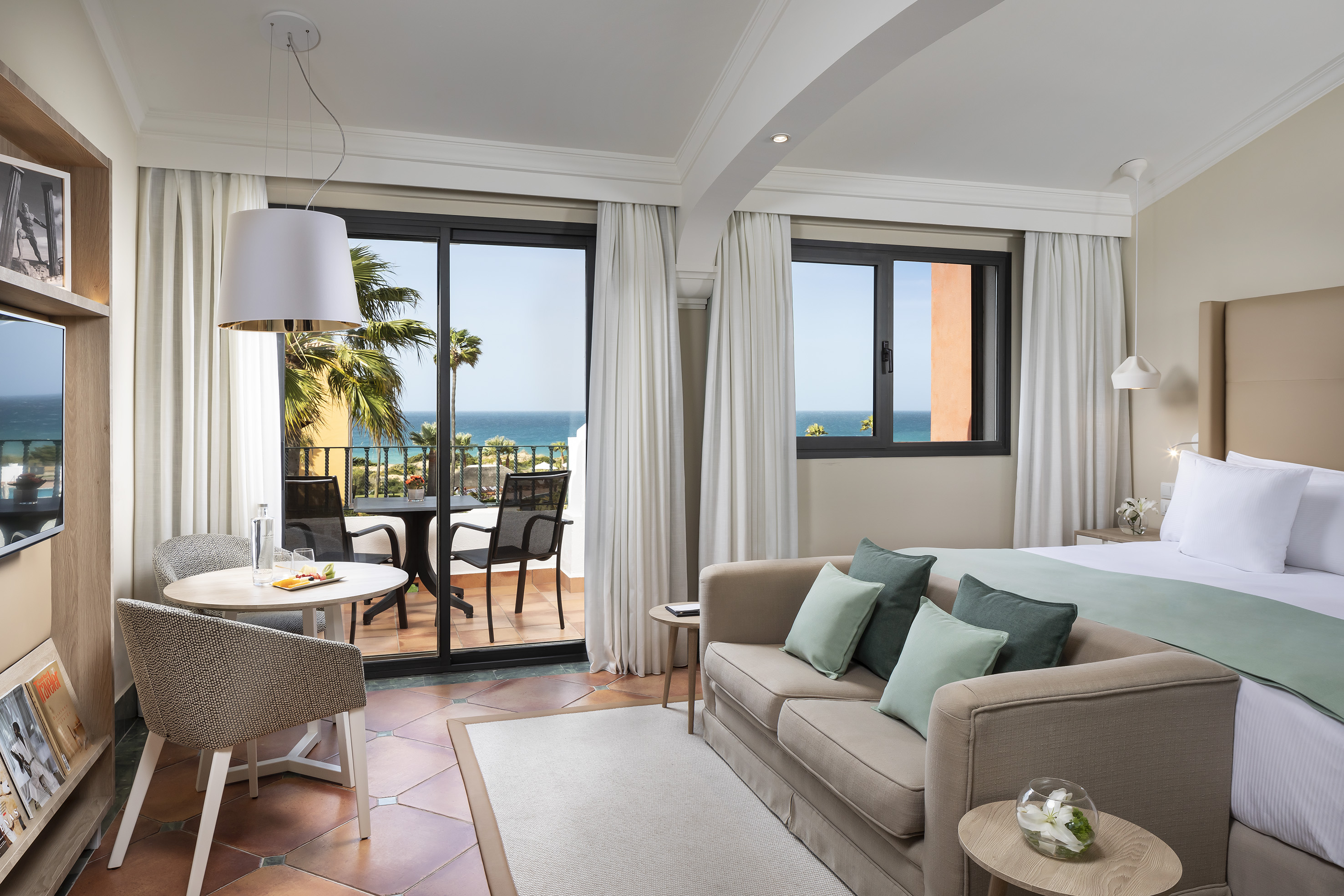 Luxury hotel suite with ocean view balcony, bed, sofa, and dining table.