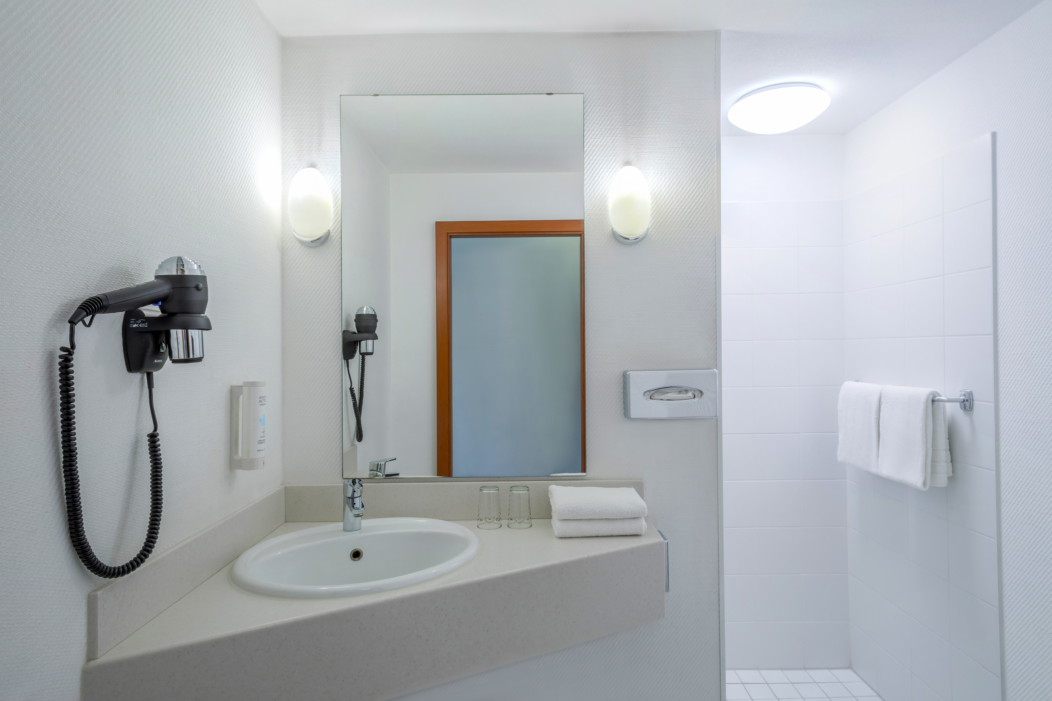 Modern hotel bathroom vanity with sink, mirror, hairdryer, and shower area.