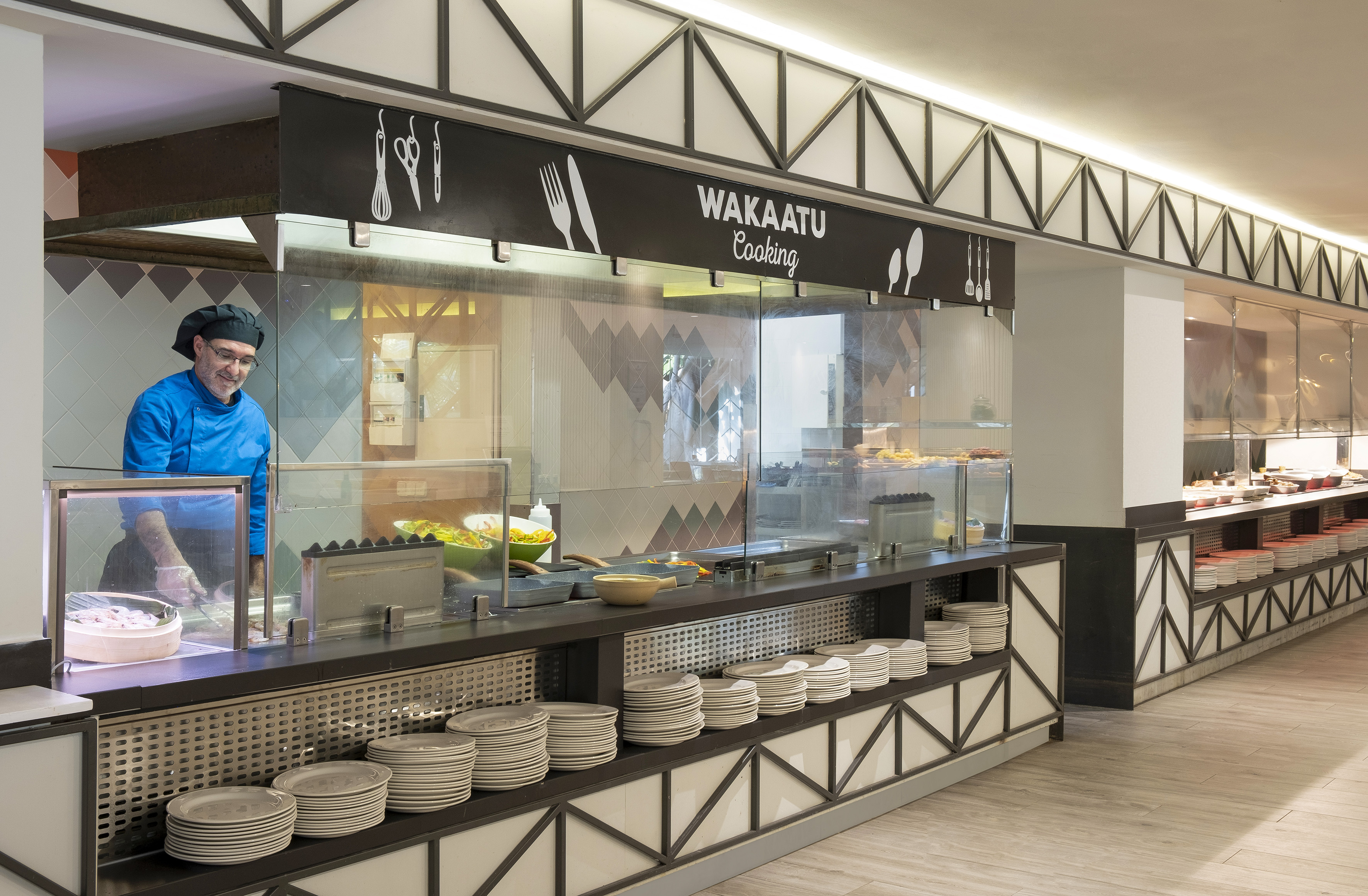 Chef in blue uniform behind Wakaatu Cooking buffet, stacked plates ready.
