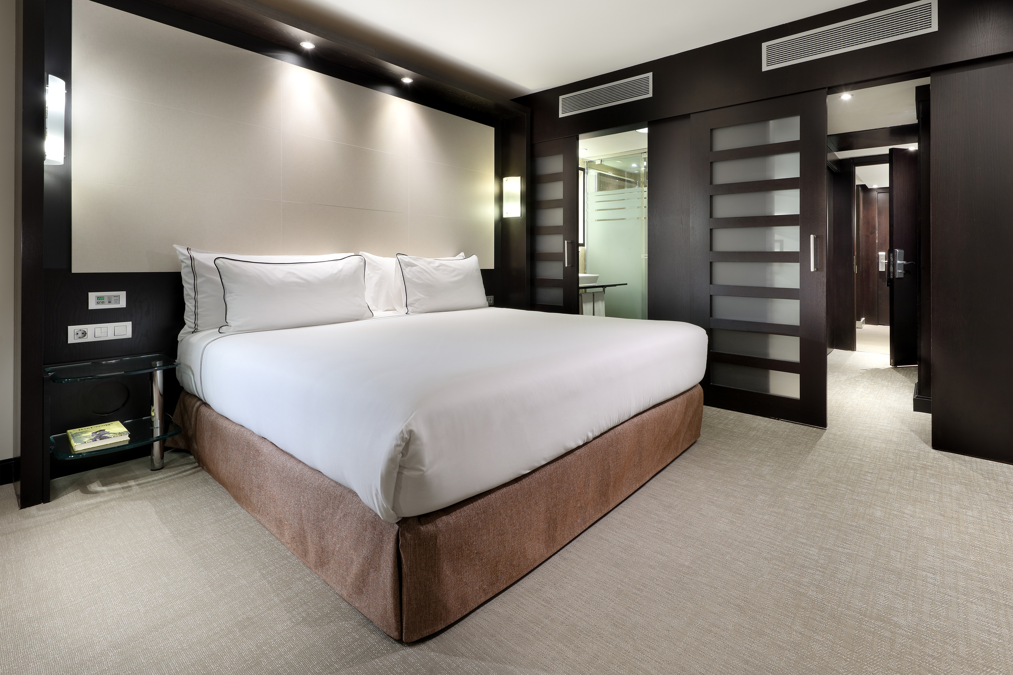Modern hotel bedroom with large white bed, dark wood decor, and en-suite bathroom.