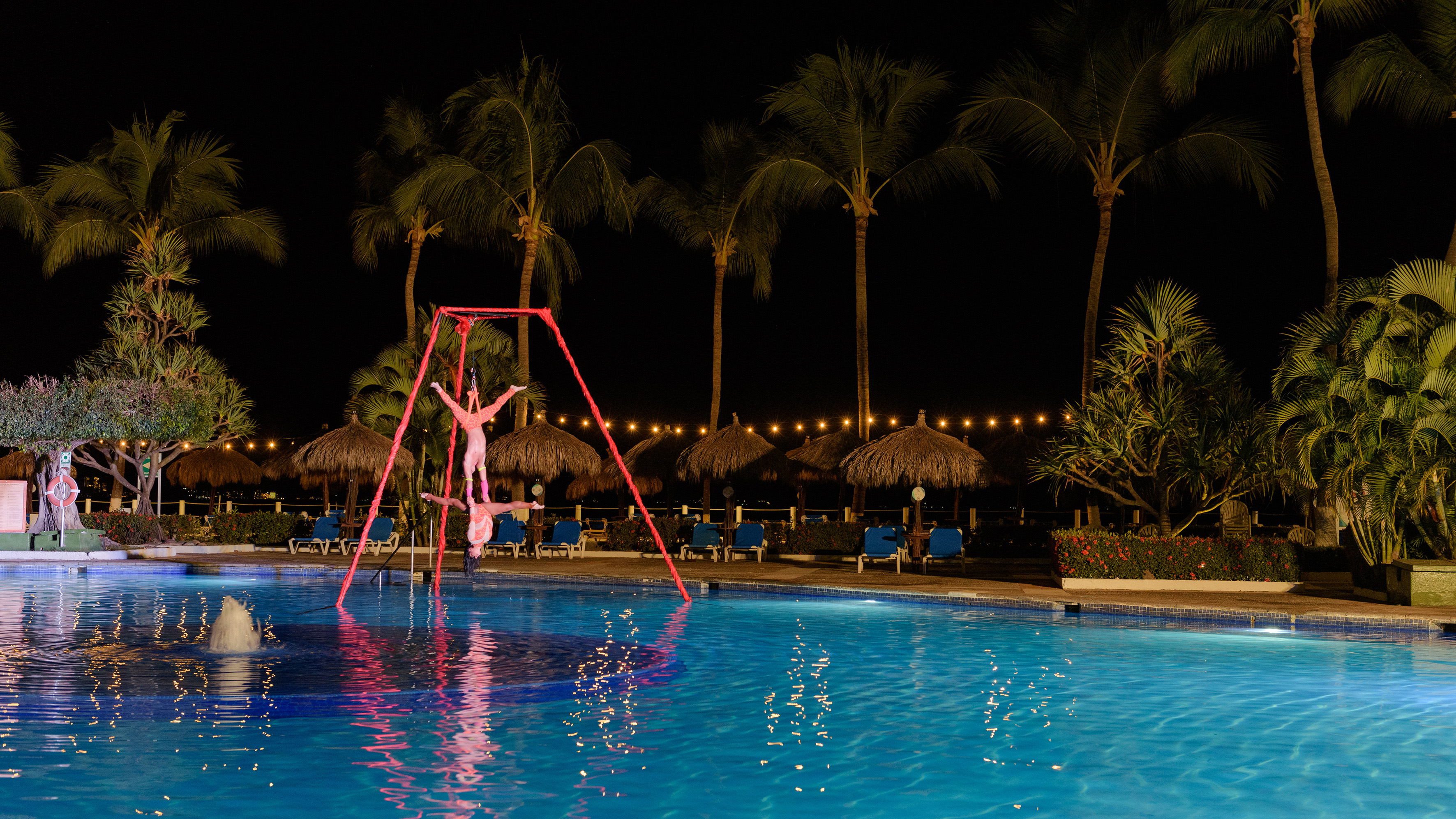 Acrobatic aerialists performing over a resort pool at night, palm trees and lights.