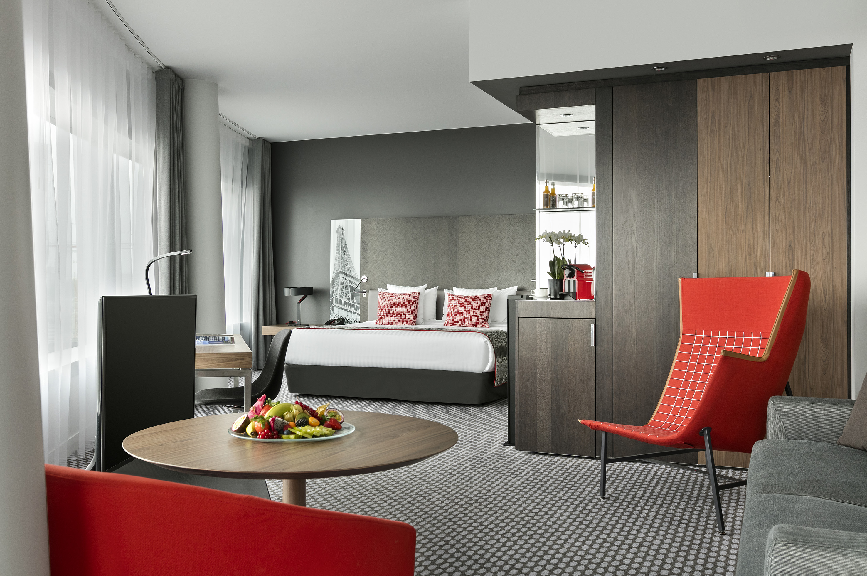 Modern hotel suite with king bed, desk, minibar, fresh fruit, and red accent chair.