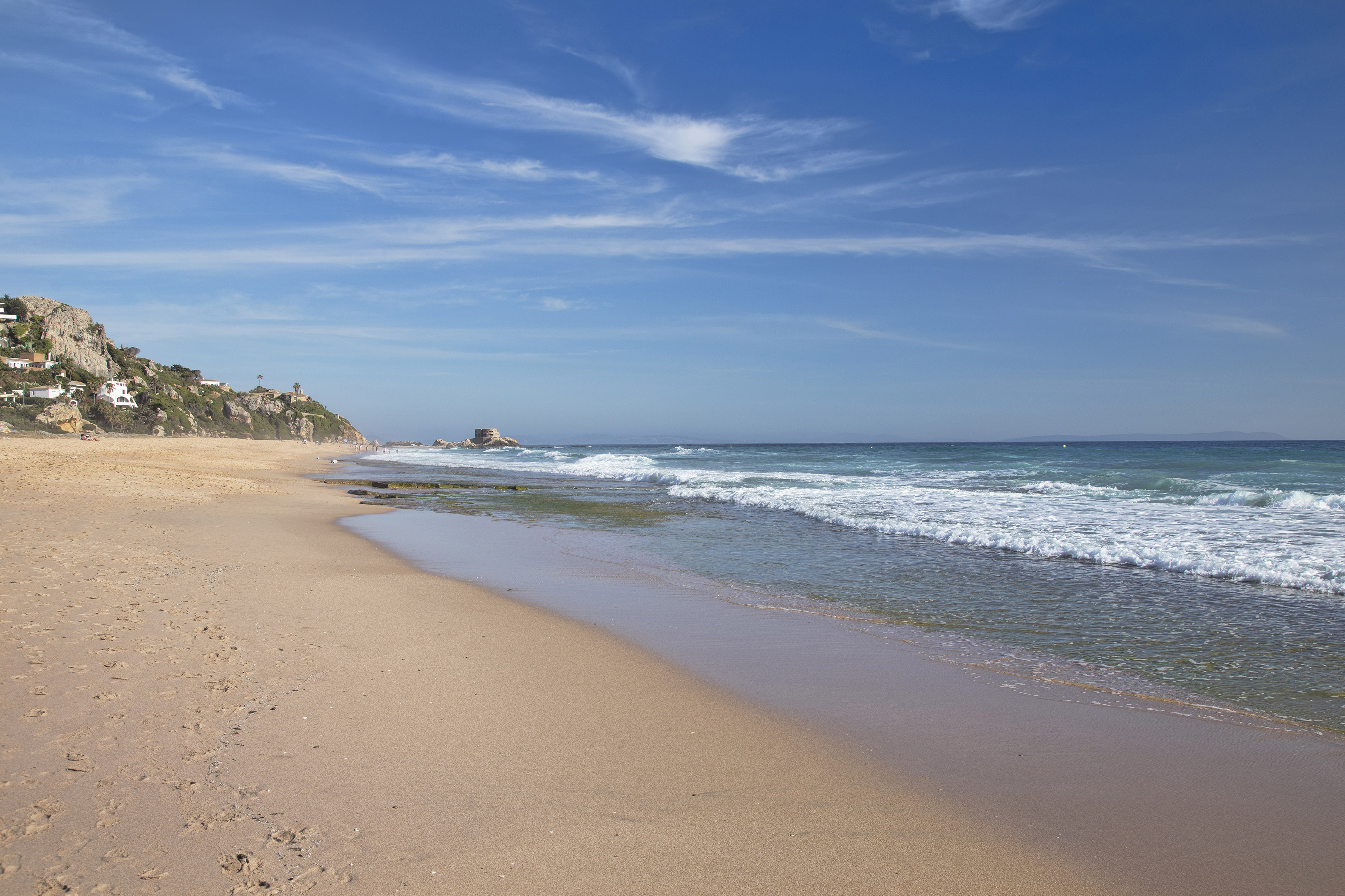 Sunny sandy beach, blue ocean waves, white hillside homes, and coastal fort.