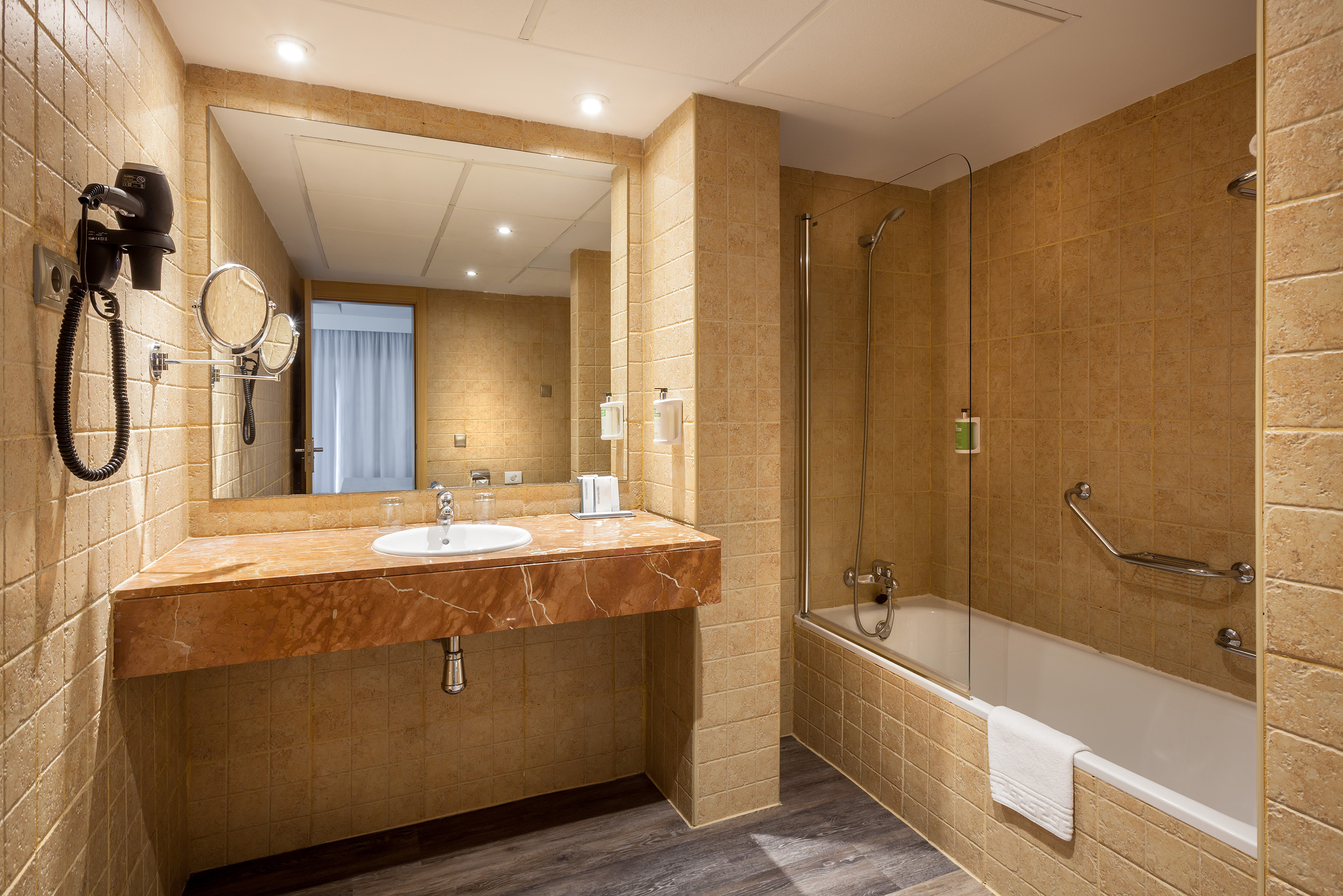 Tiled hotel bathroom with vanity, large mirror, and bathtub-shower.