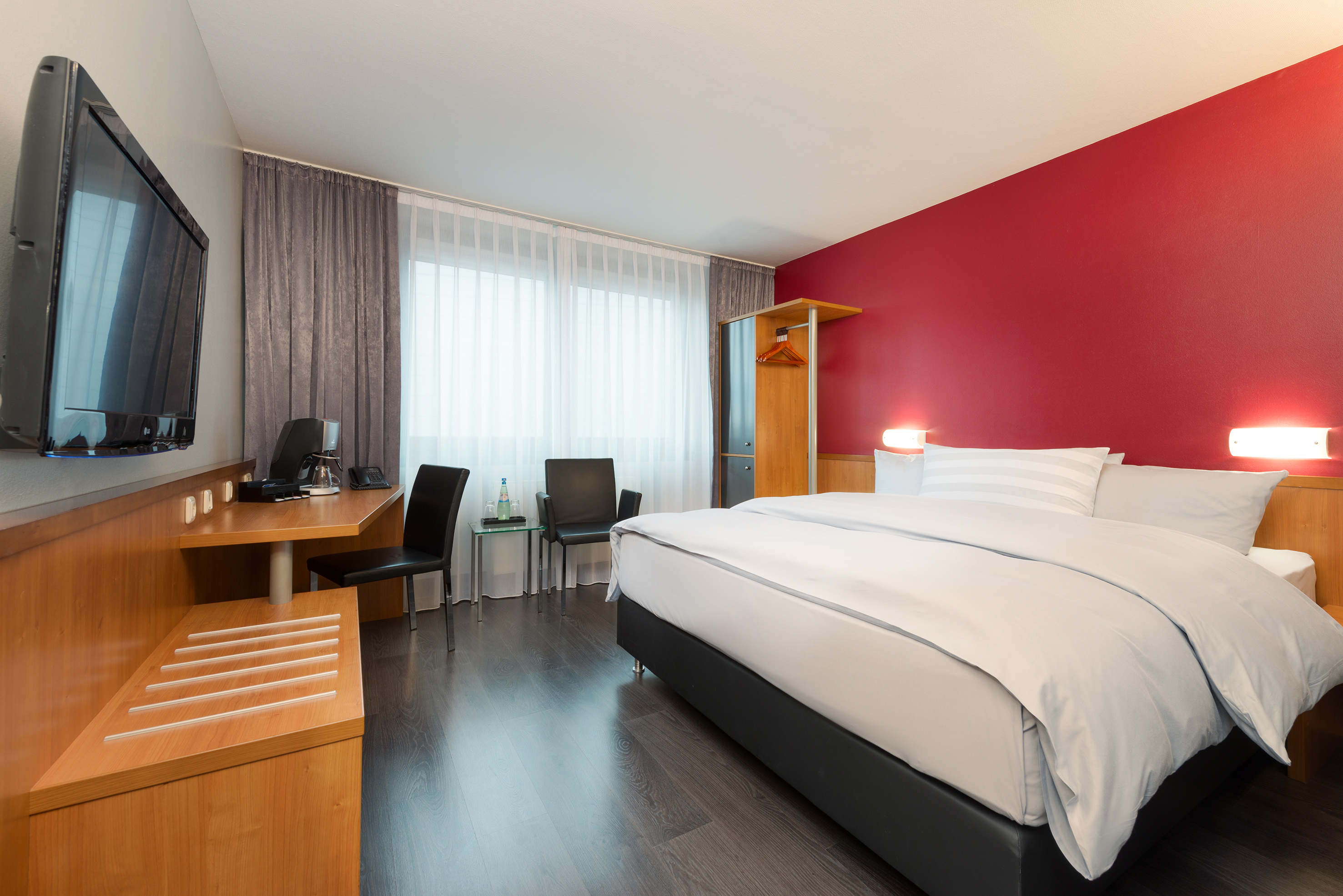 Hotel bedroom with queen bed, red accent wall, dark wood floor, desk, and TV.