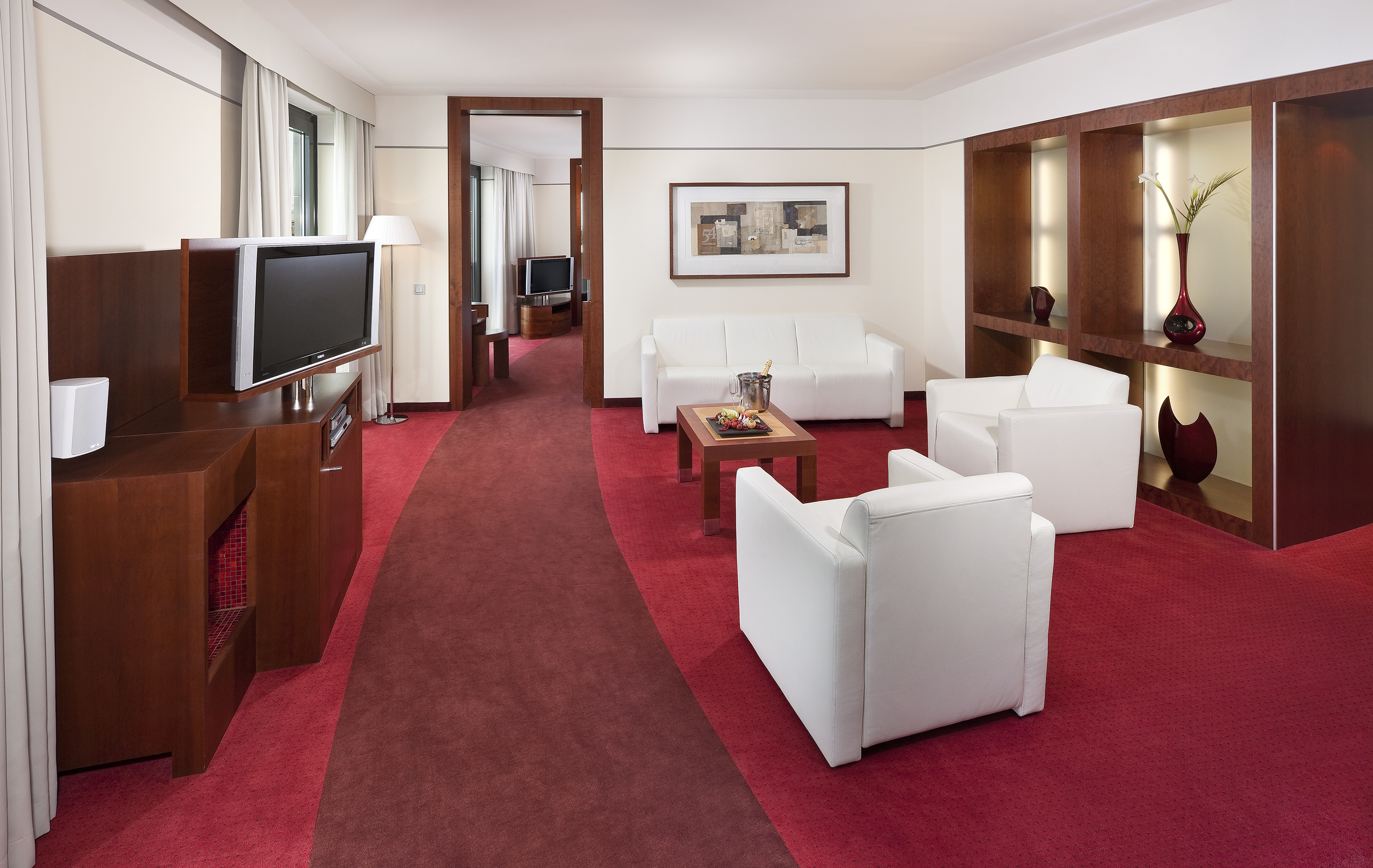 Modern hotel suite living room with white sofas, red carpet, and wooden accents.
