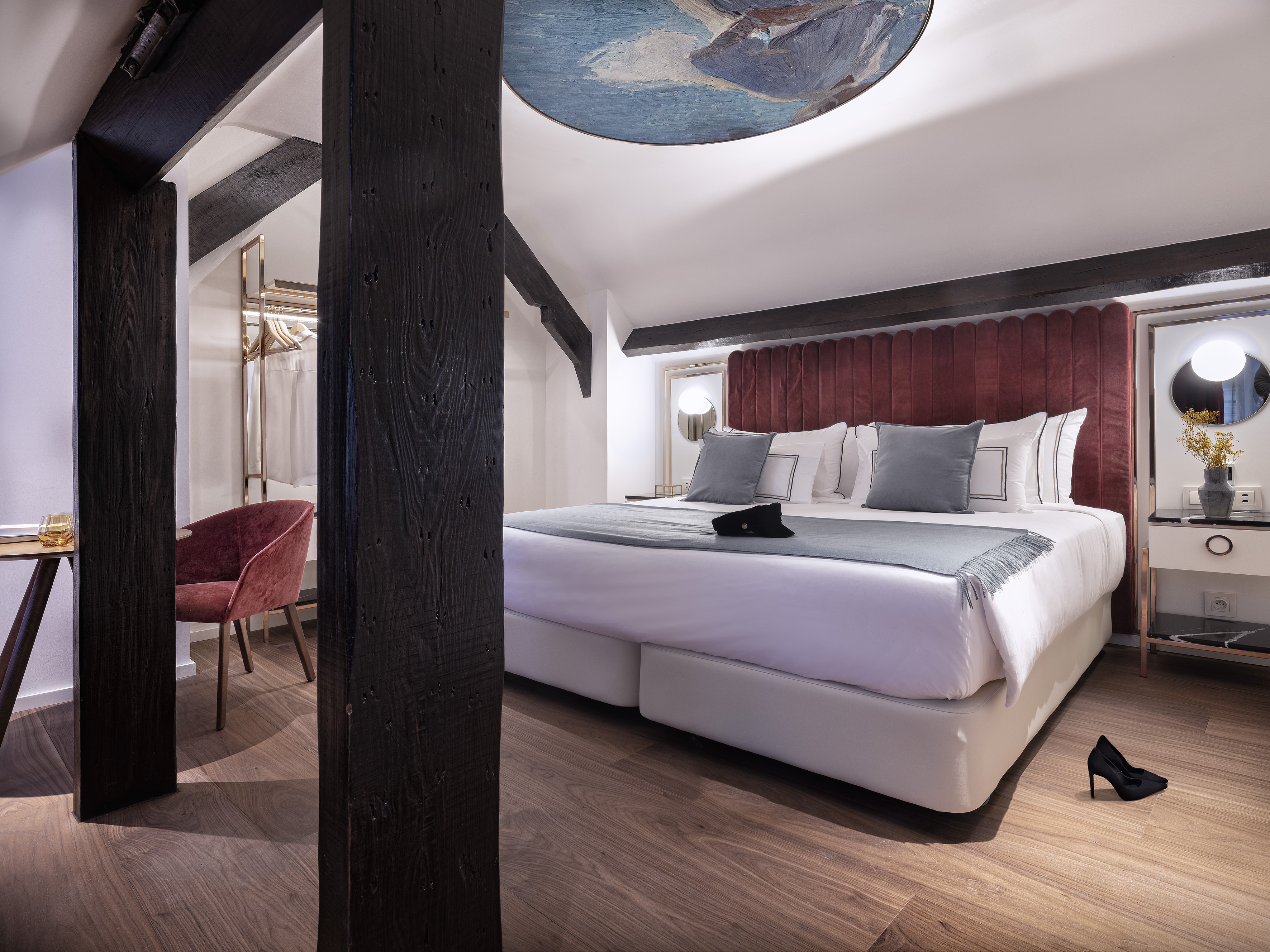 Luxury hotel bedroom with velvet bed, exposed wood beams, and ceiling art.