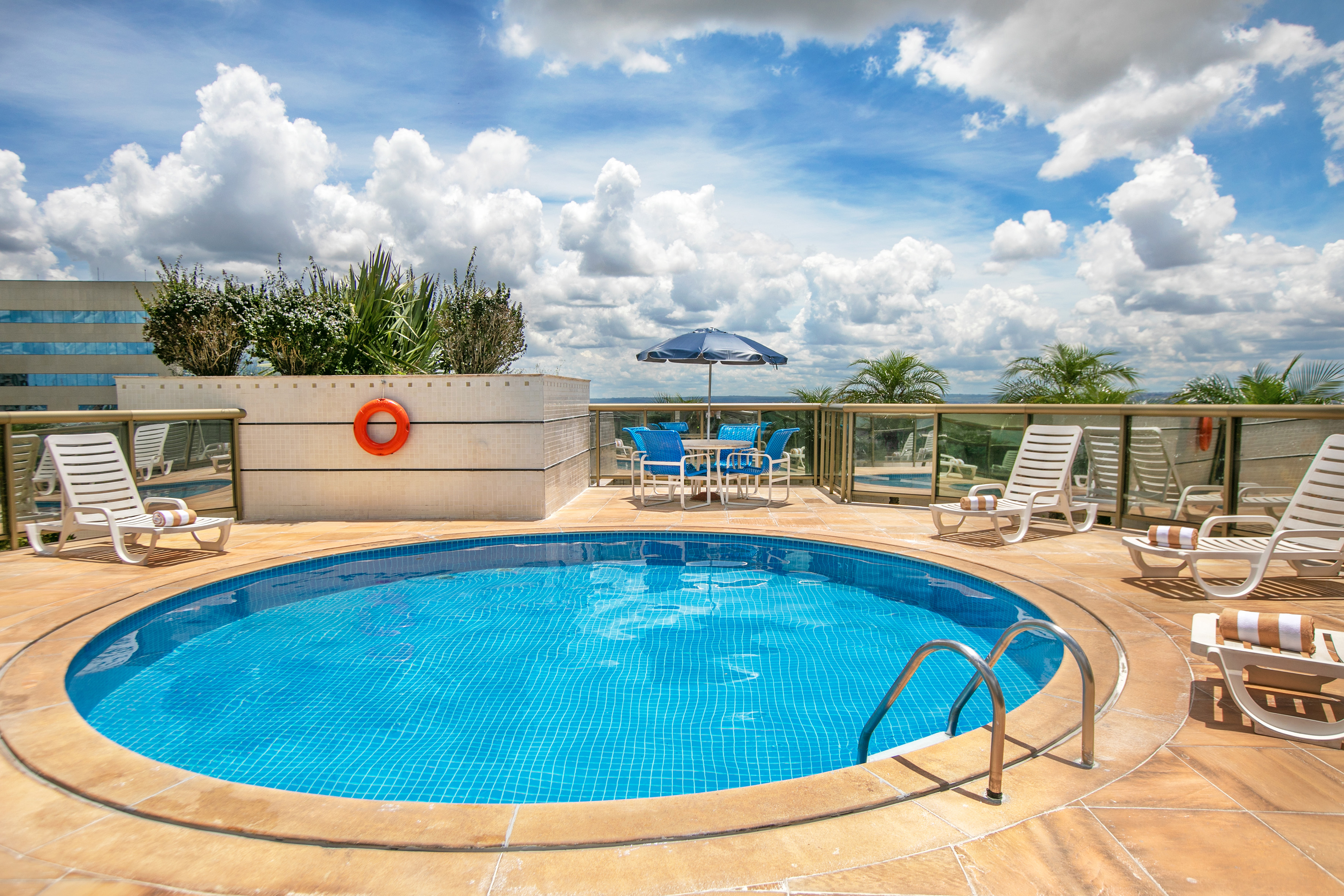 Sunny hotel rooftop pool with blue tiled water, chairs, and umbrella.