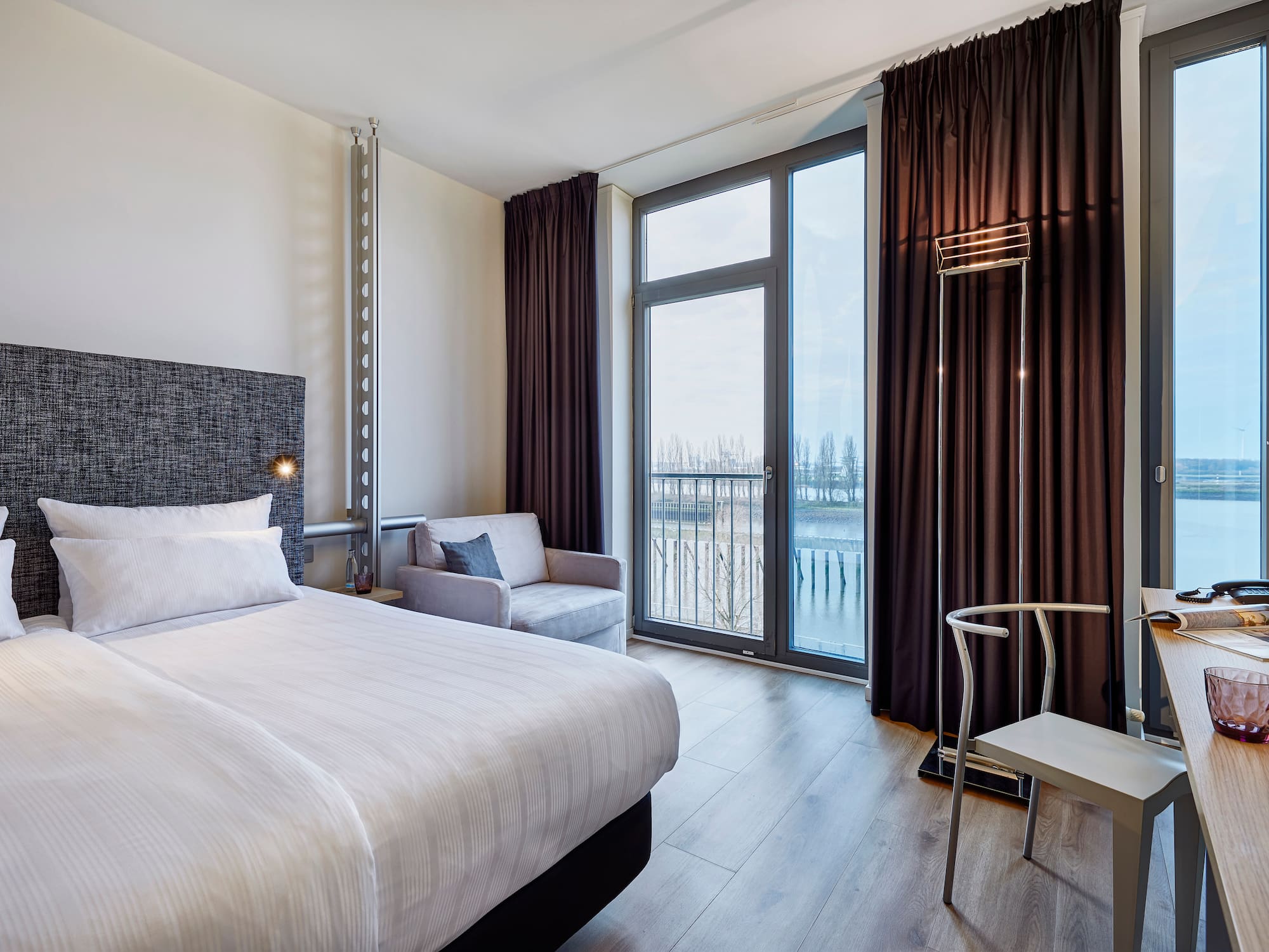 Modern hotel room with bed, desk, armchair, and river view from large windows.