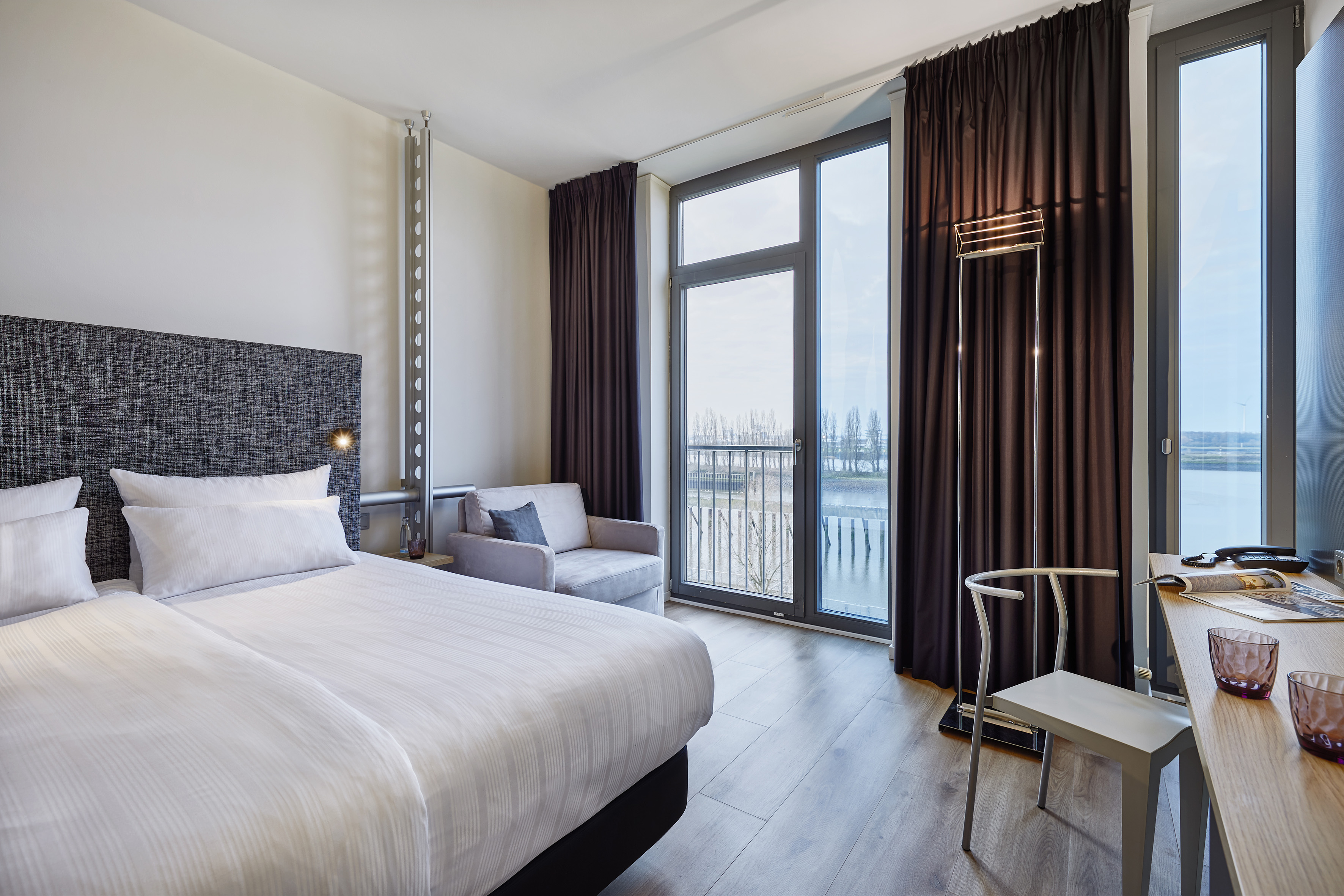Modern hotel room with bed, desk, armchair, and river view from large windows.
