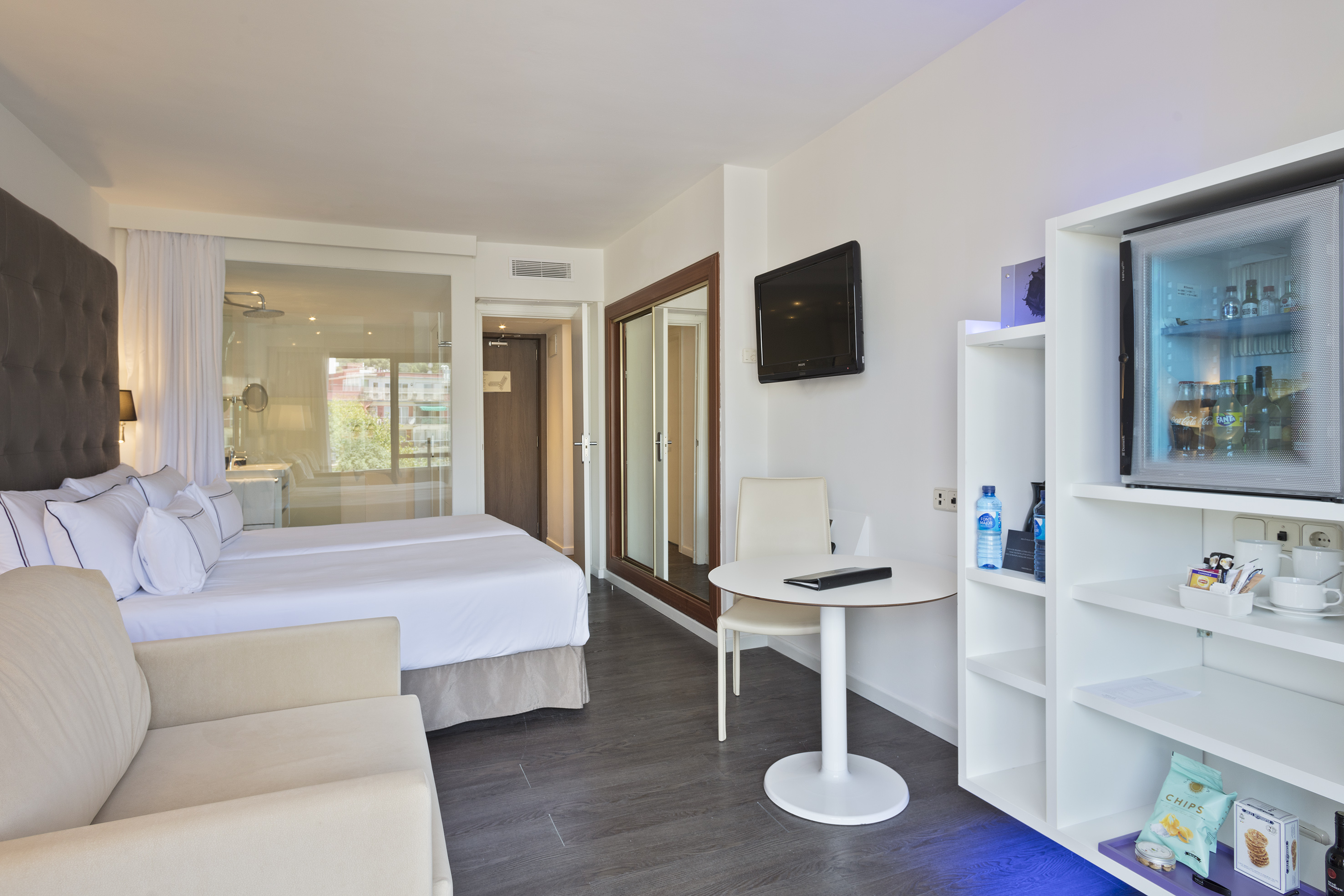 Bright hotel room with twin beds, TV, mini-bar, and glass-partitioned bathroom.