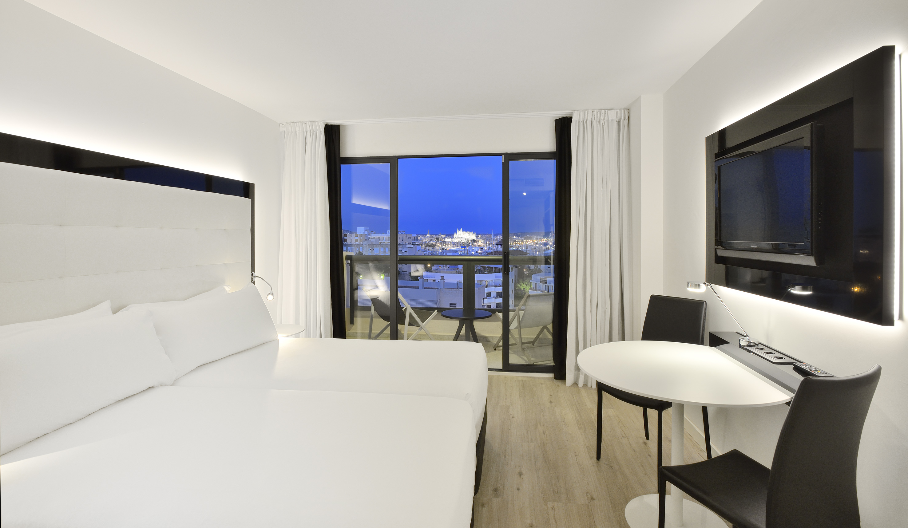 Modern hotel room with city night view balcony, white bed, flat-screen TV.