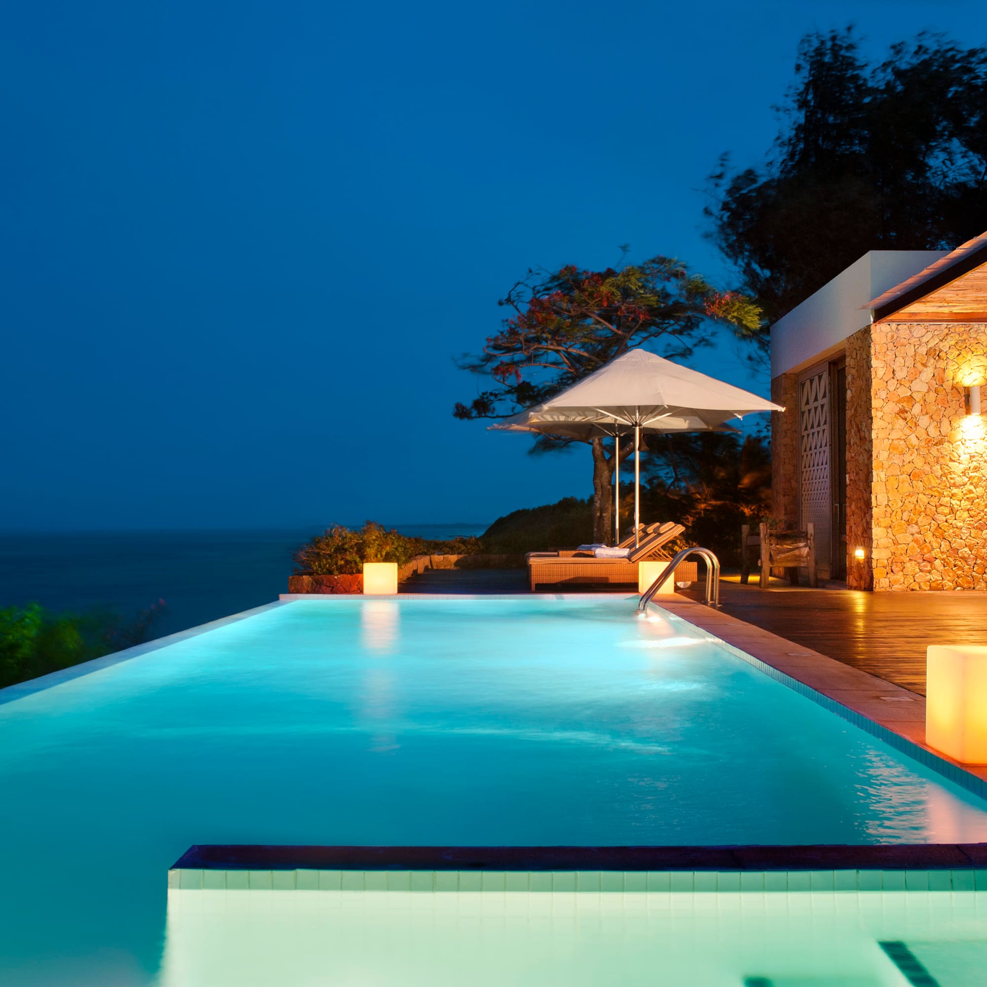 a pool with a deck and umbrella and a building with trees and a blue sky