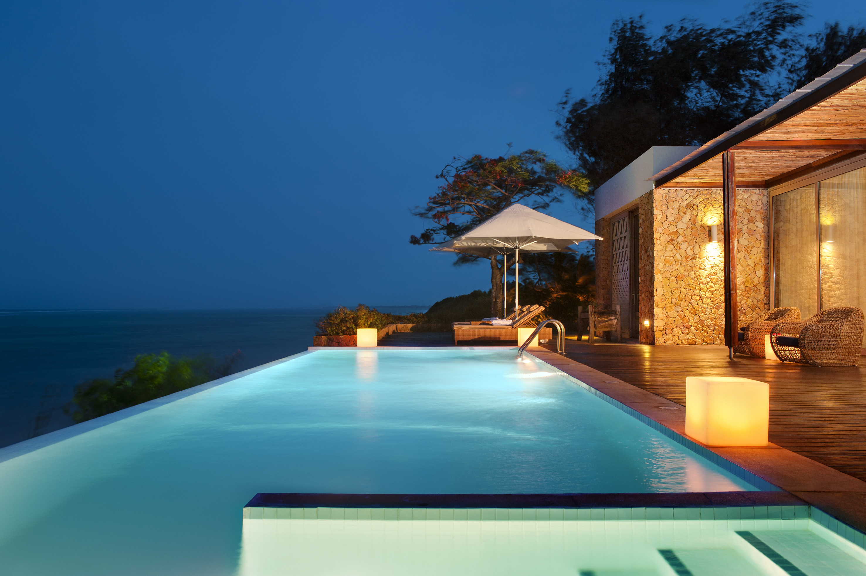 Tropical resort infinity pool with ocean view and illuminated villa at night.