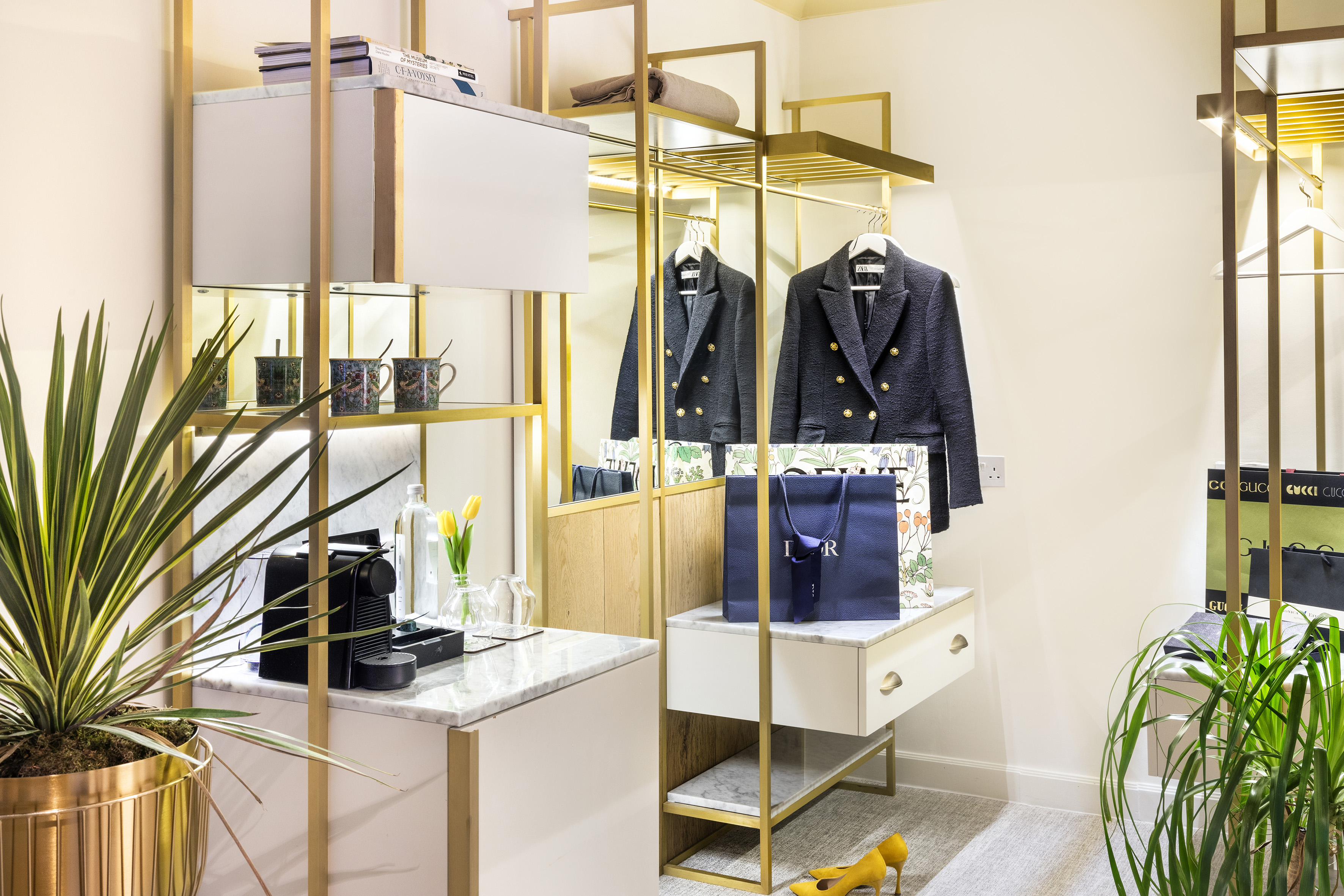 Hotel room wardrobe with gold frame, marble bar, blazers, coffee machine, and plants.