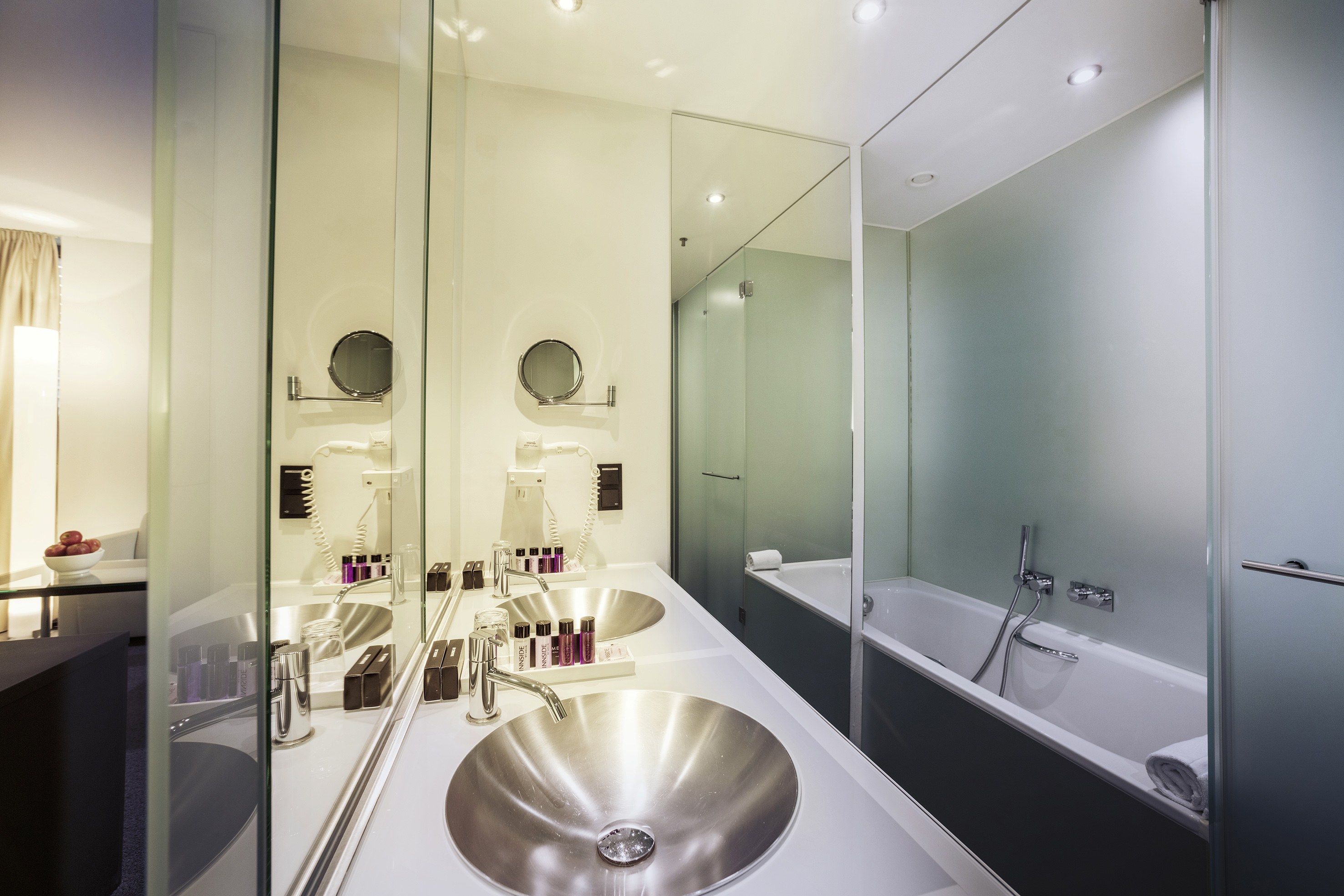 Sleek hotel bathroom with double vanity, metallic sinks, and glass tub enclosure.