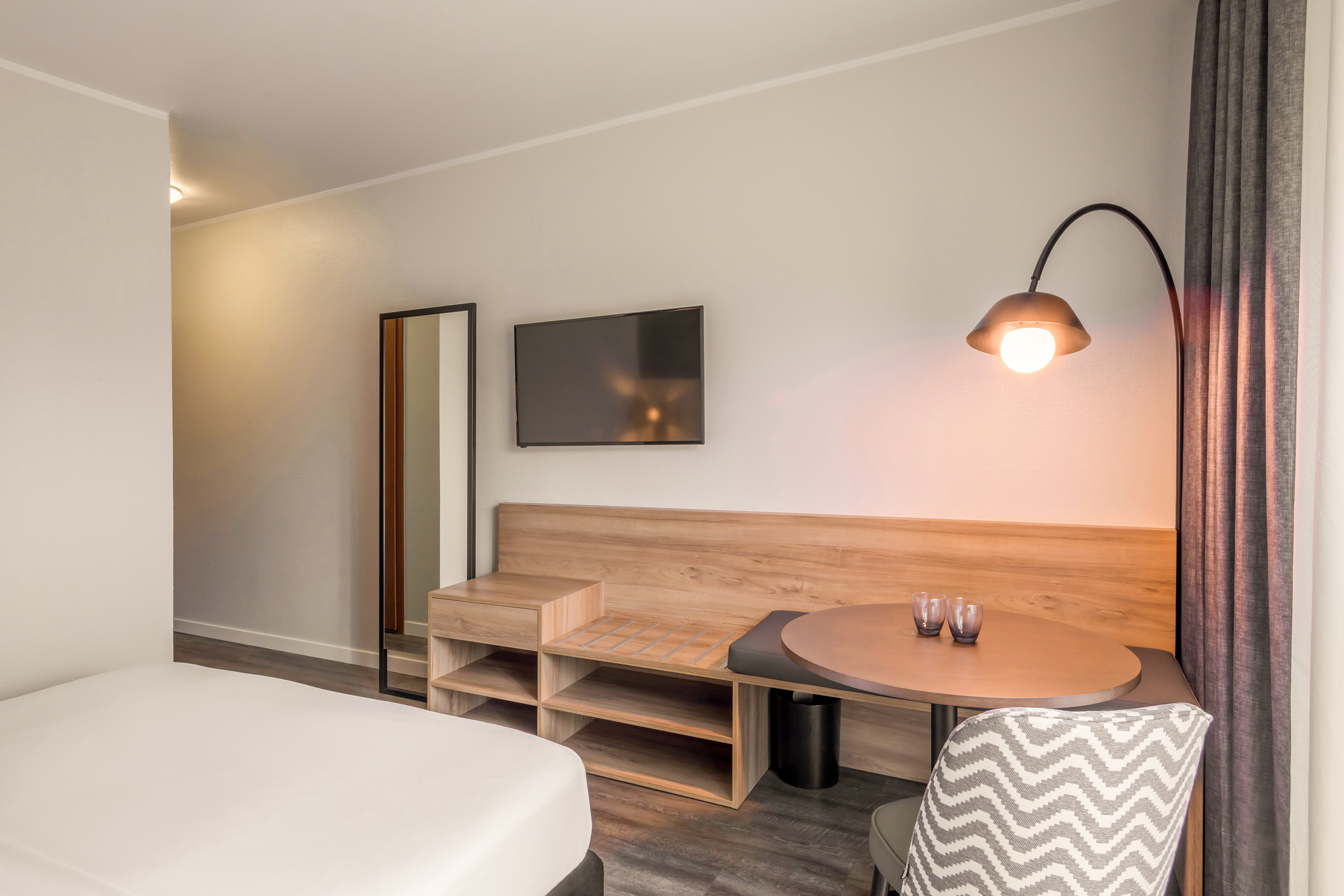 Modern hotel room with bed, TV, wooden storage, dining table, and contemporary lamp.