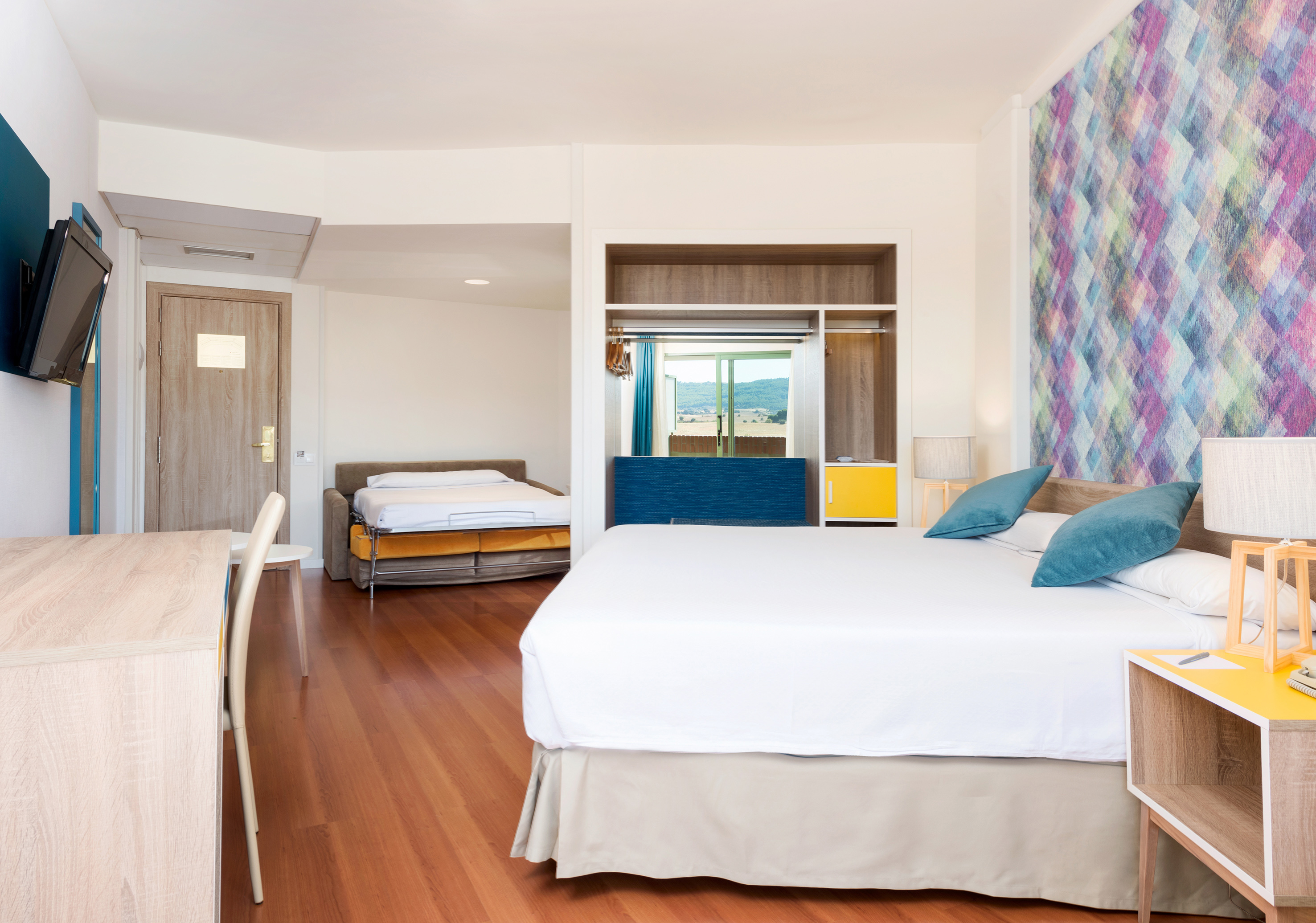 Modern hotel room with large bed, colorful accent wall, desk, and sofa bed.