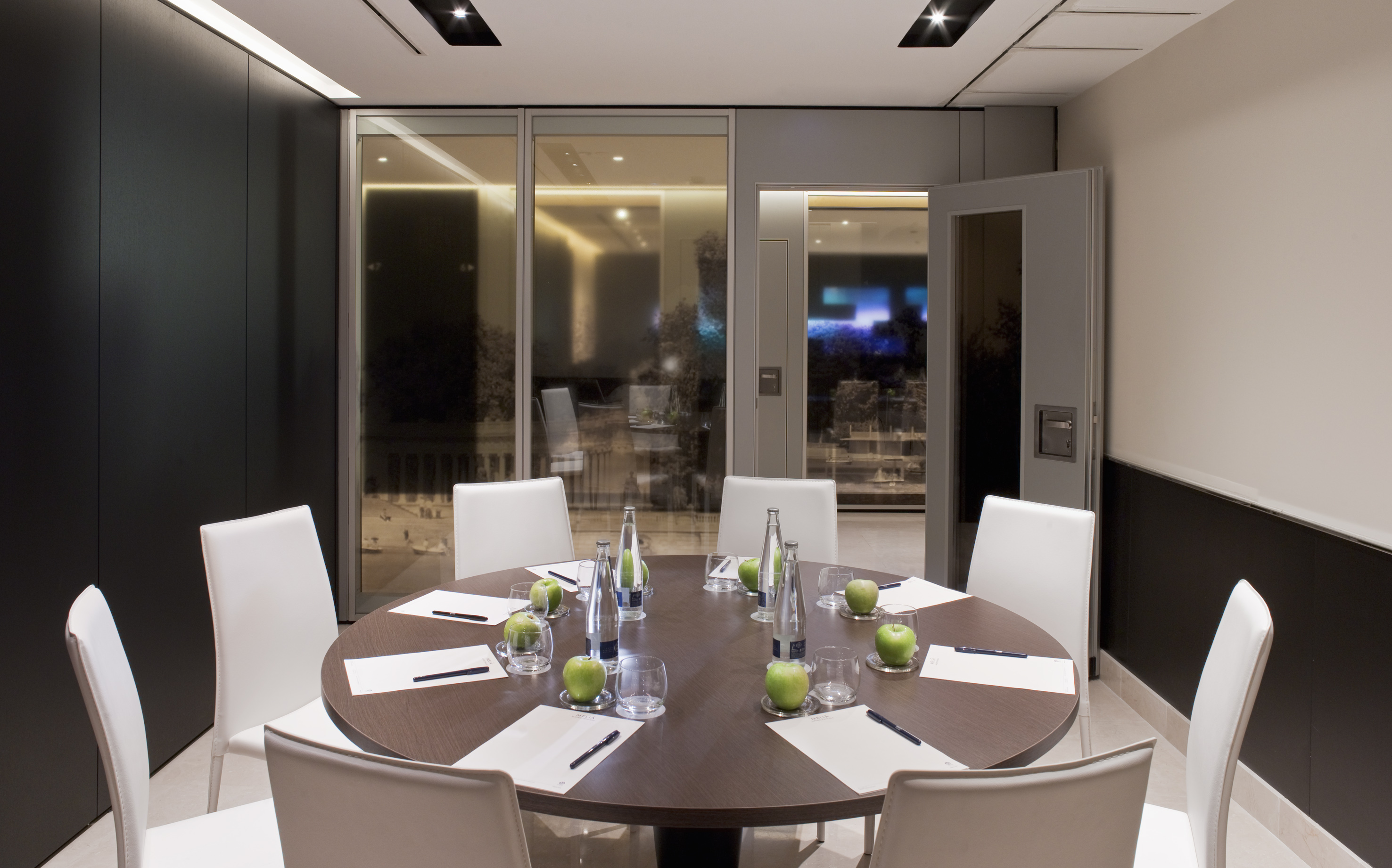 Modern meeting room with round wood table, white chairs, bottled water, and apples.