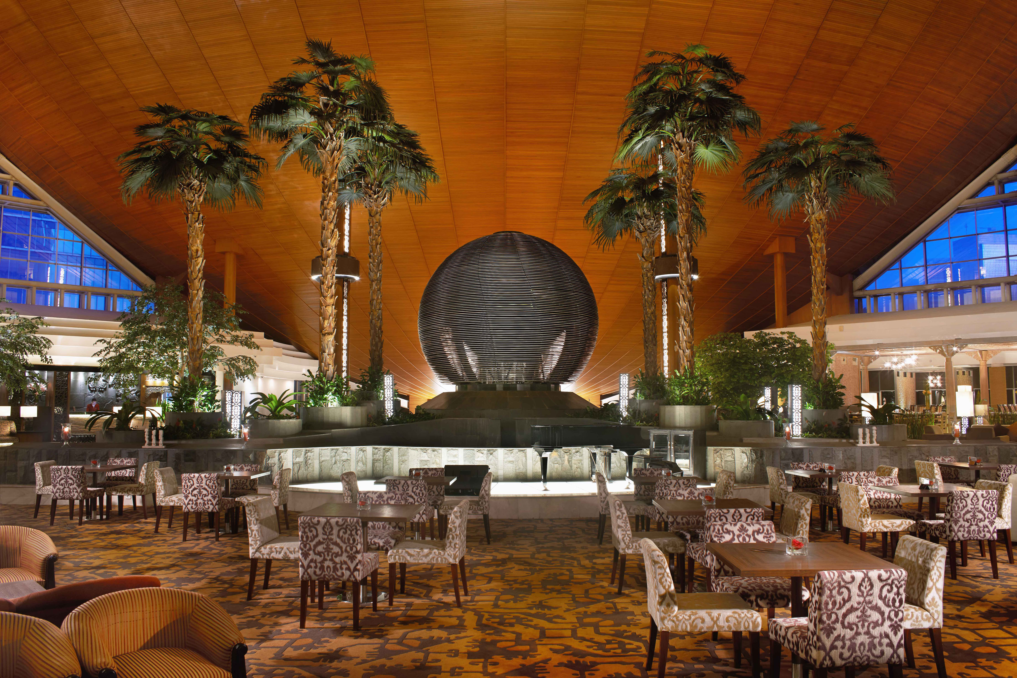 Grand hotel lobby with palm trees, sphere art, and patterned lounge seating.