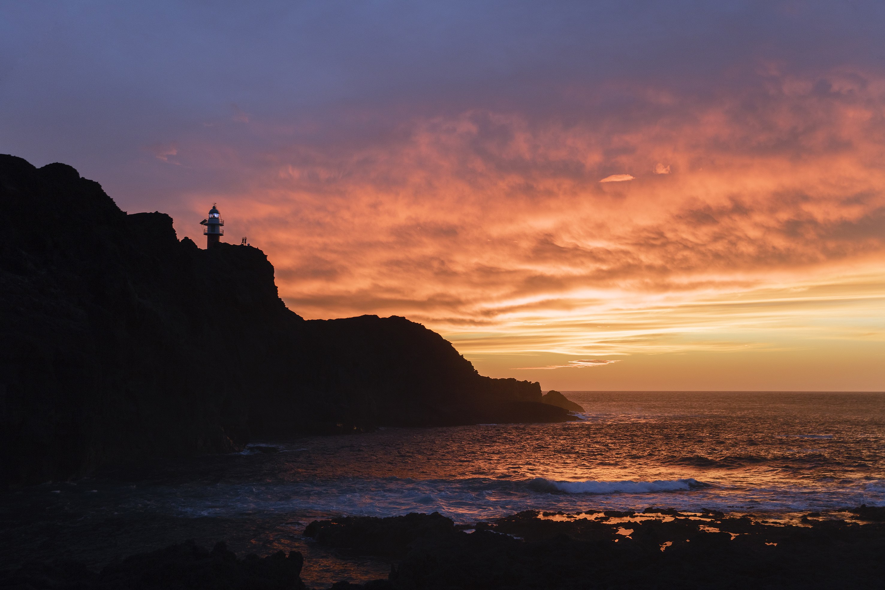 Coastal lighthouse on cliff overlooking ocean at vivid sunset.