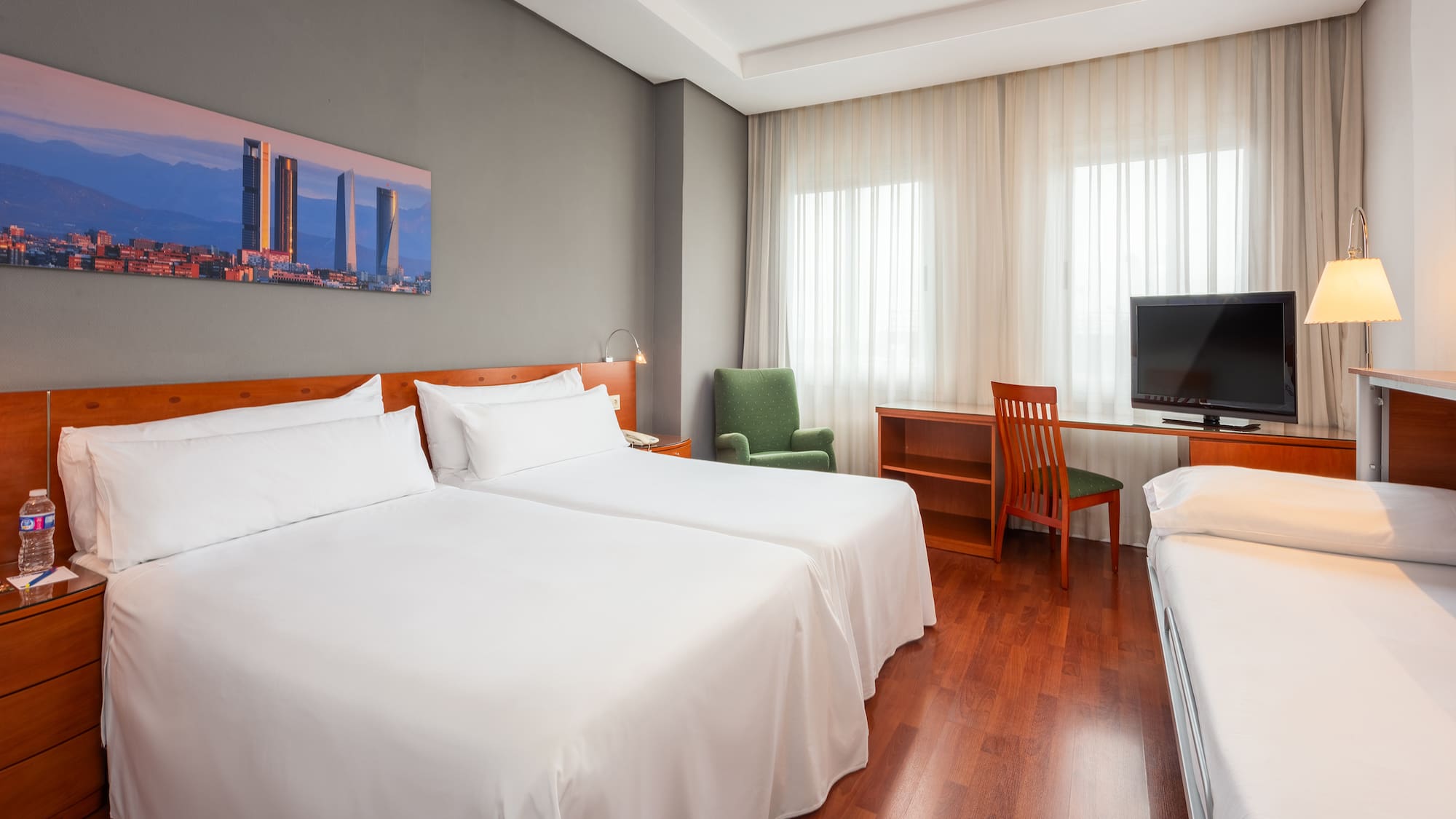 Hotel family room with twin beds, extra bed, desk, TV, and city art.