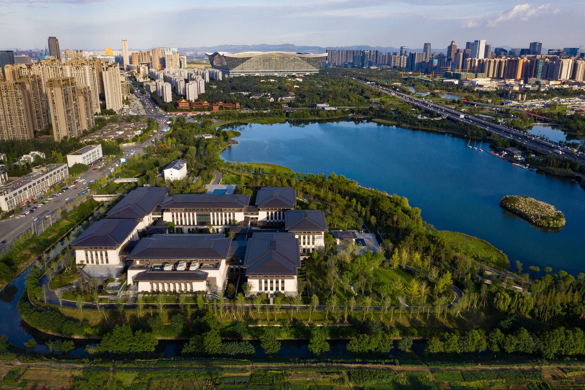 Aerial city view: Modern lakeside buildings, green parks, highway, and urban skyline.