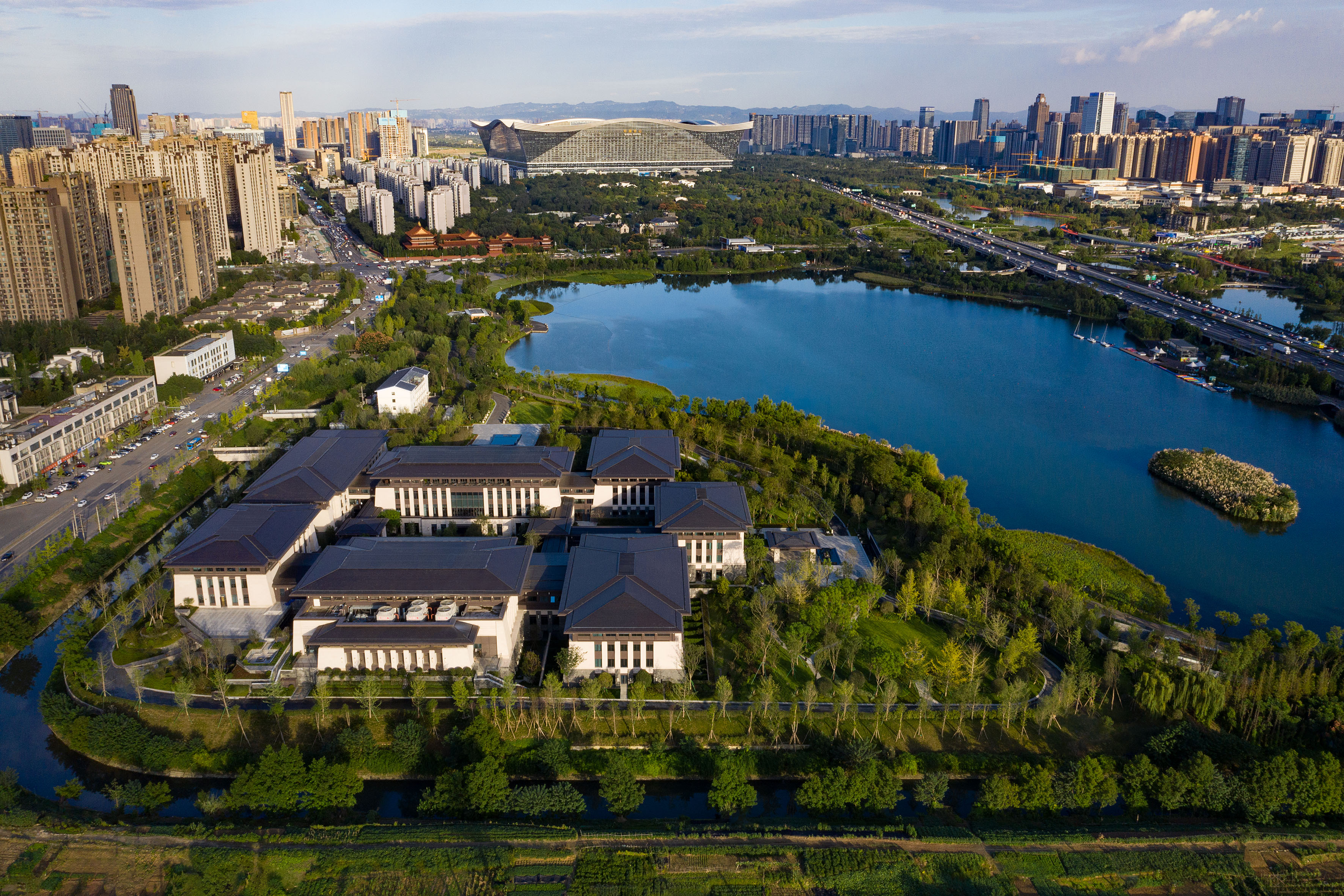 Aerial city view: Modern lakeside buildings, green parks, highway, and urban skyline.