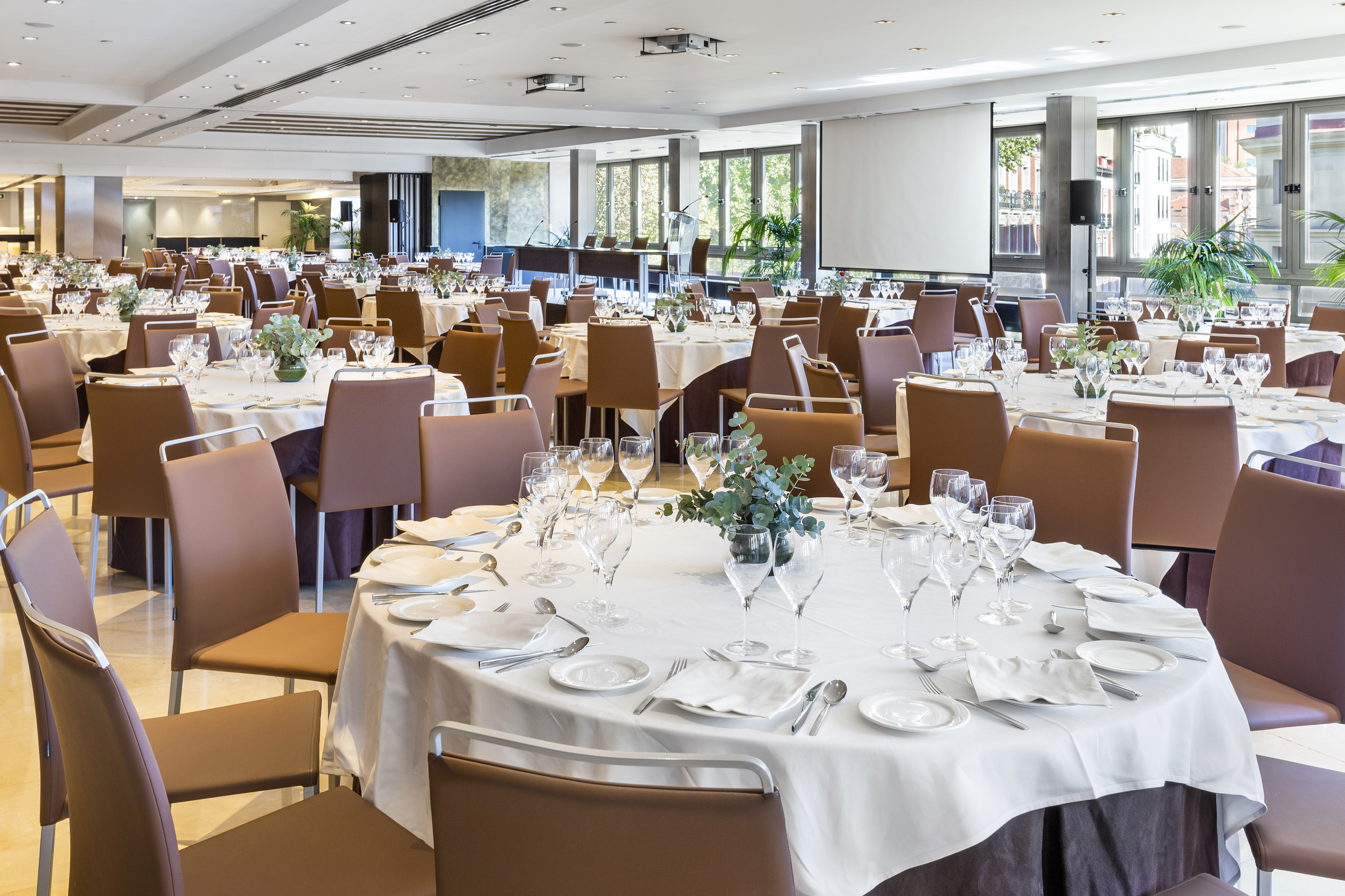 Spacious event room with set round tables, white linens, brown chairs.