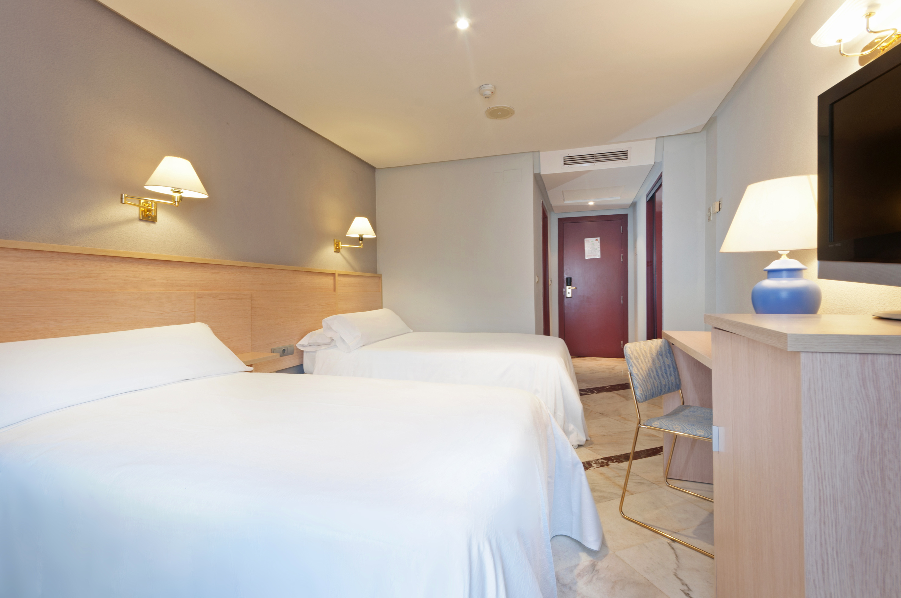 Hotel room with two twin beds, white sheets, desk with TV, and red door.