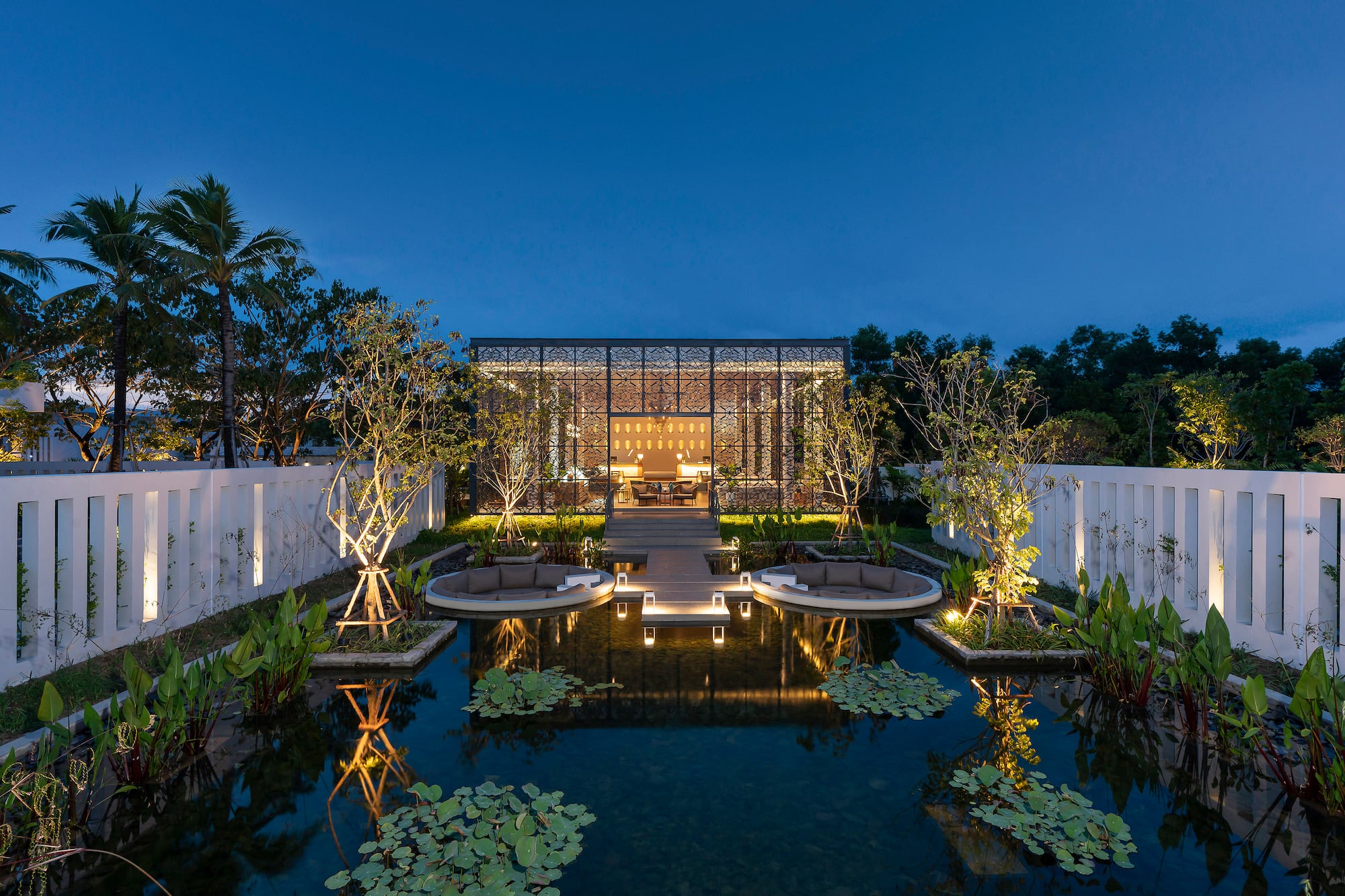 Illuminated modern pavilion, reflecting pond, and outdoor lounge seating at dusk.