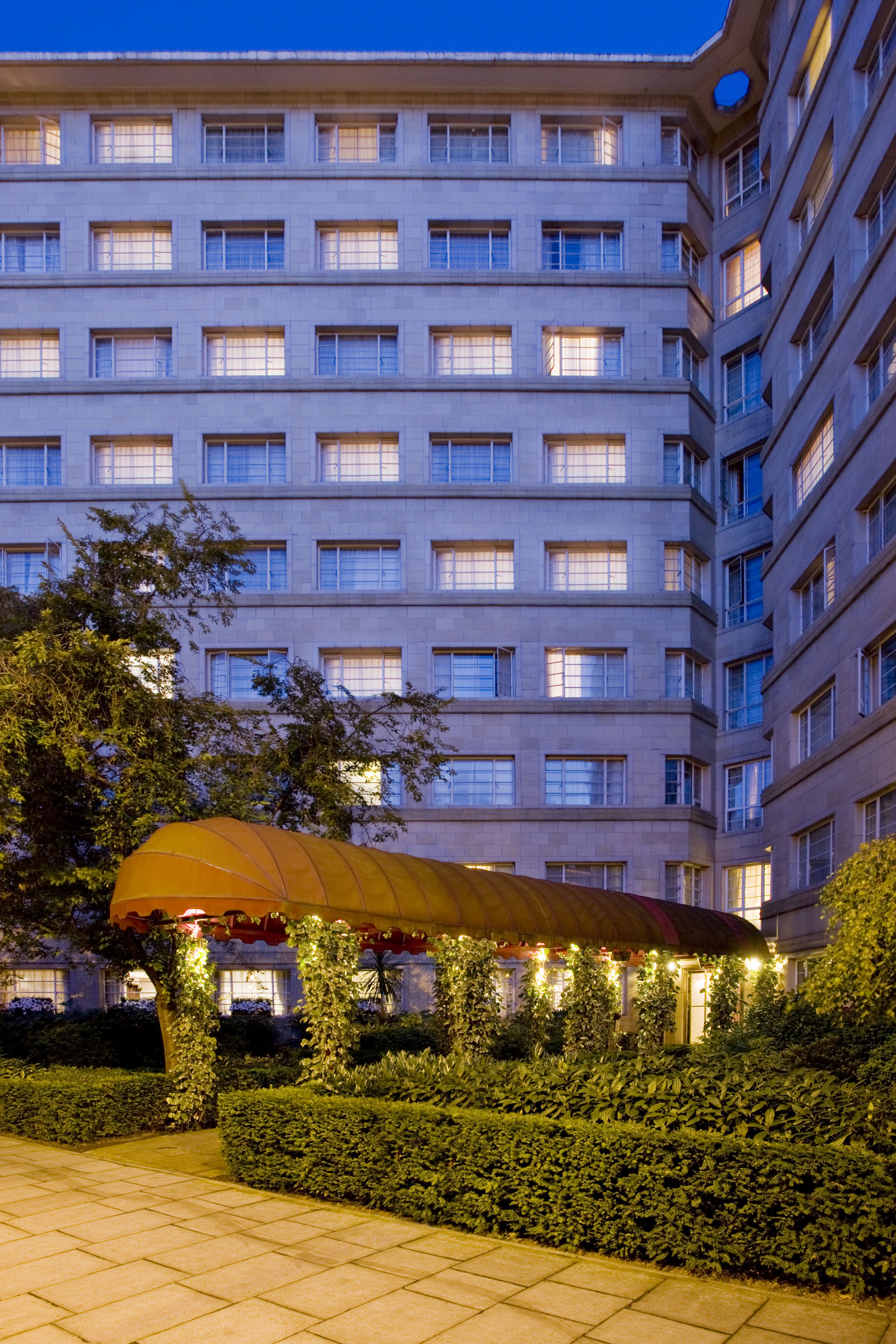 Hotel entrance at night, featuring a lit orange awning and lush garden.