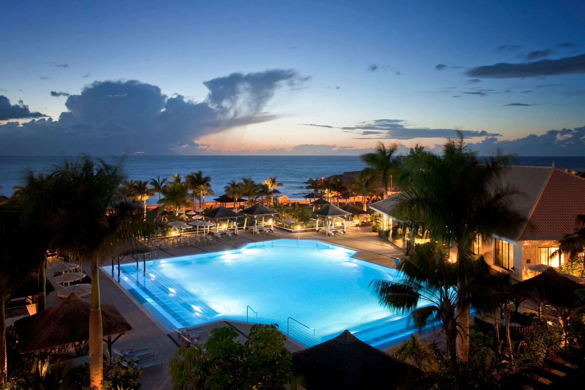 Illuminated resort swimming pool at dusk with ocean and palm trees.