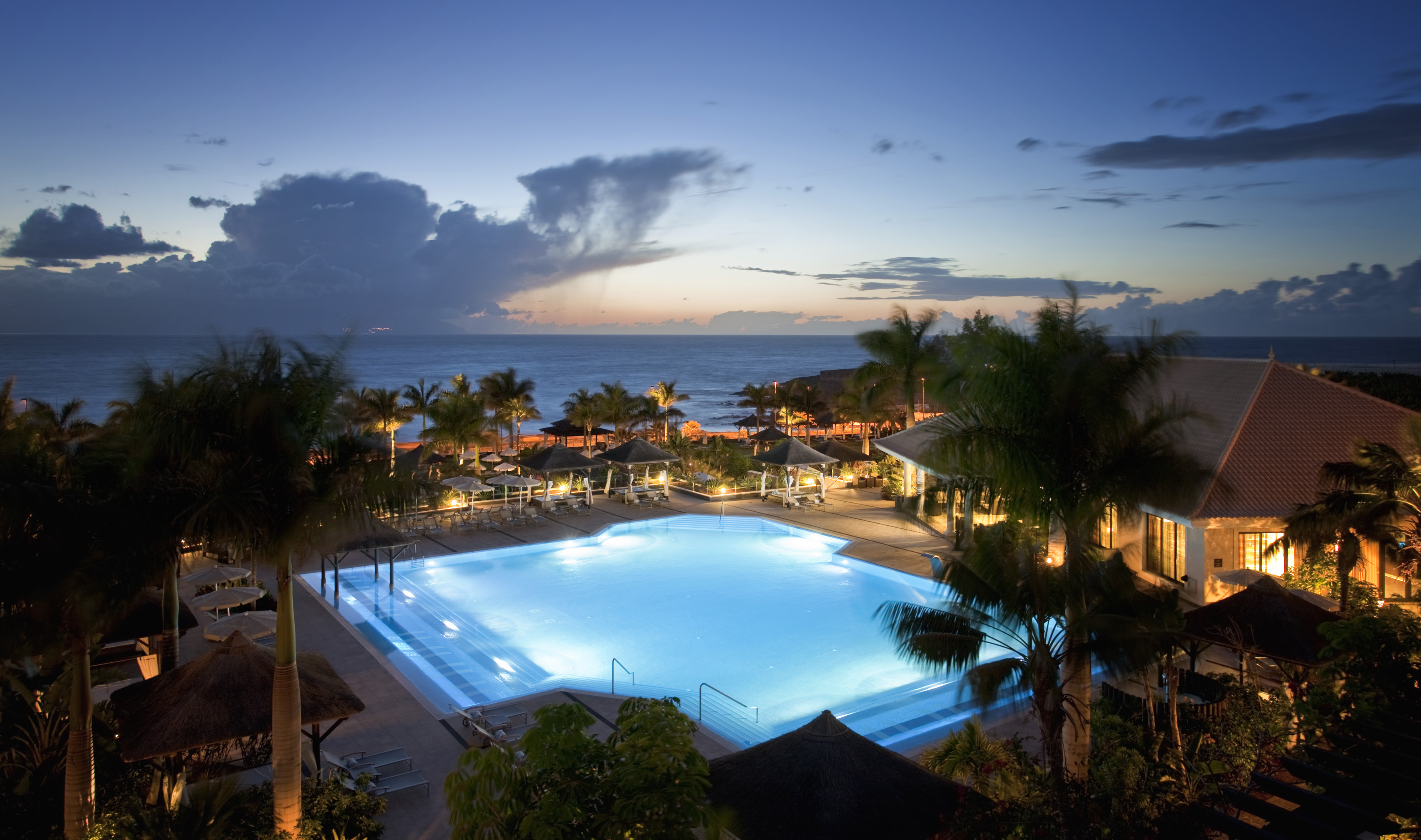 Illuminated resort swimming pool at dusk with ocean and palm trees.