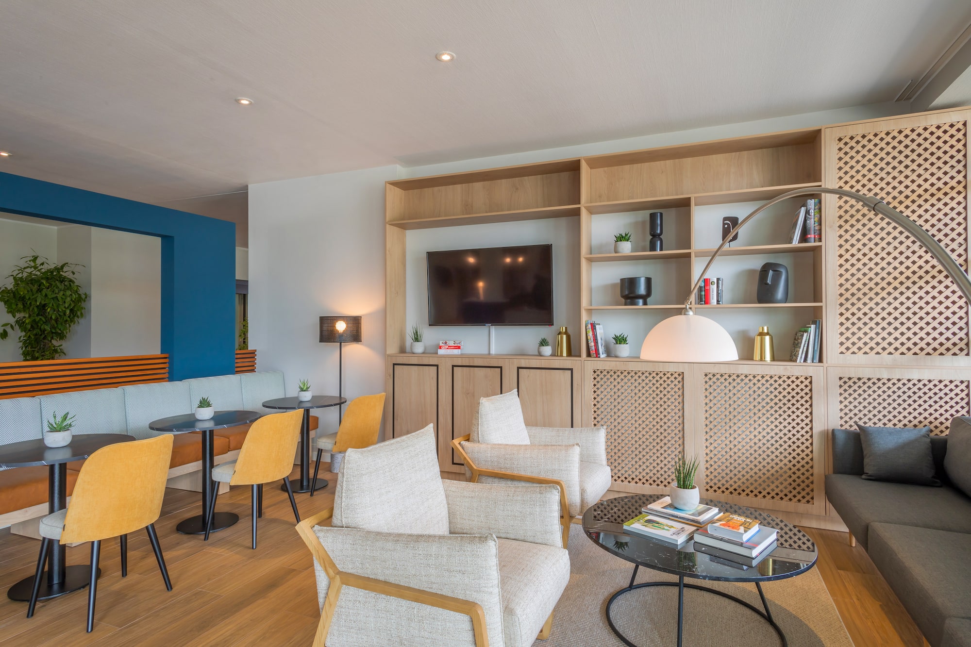 Modern hotel lounge with yellow chairs, armchairs, TV, and wooden shelving.
