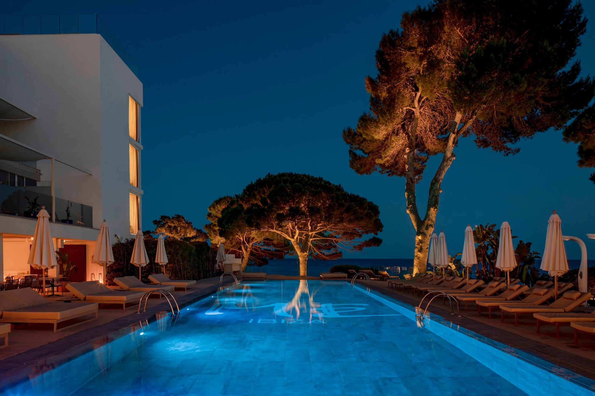 Illuminated Ibiza resort pool at night with loungers, trees, and ocean.