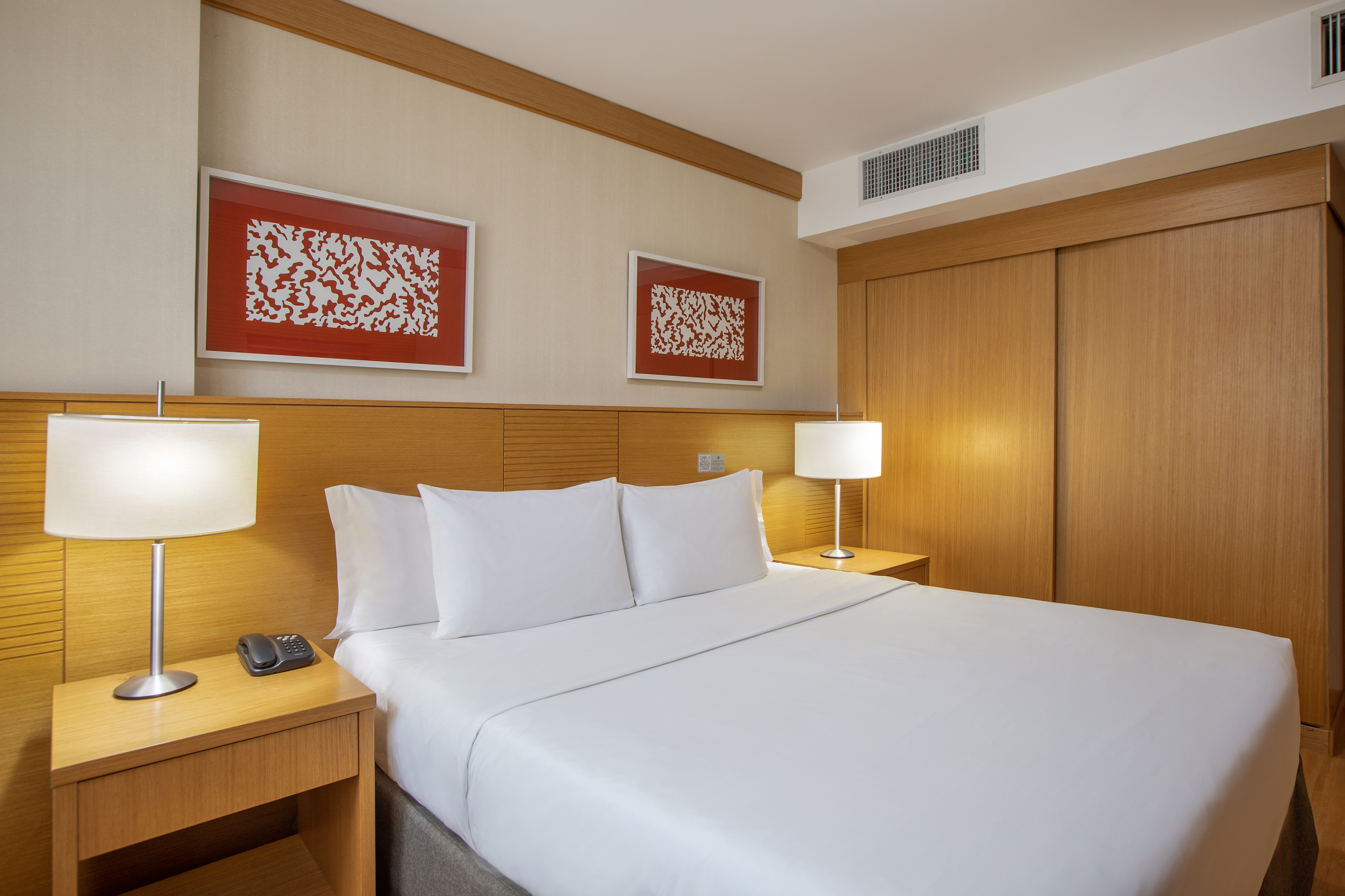 Modern hotel room with queen bed, white linens, wooden nightstands, red art.