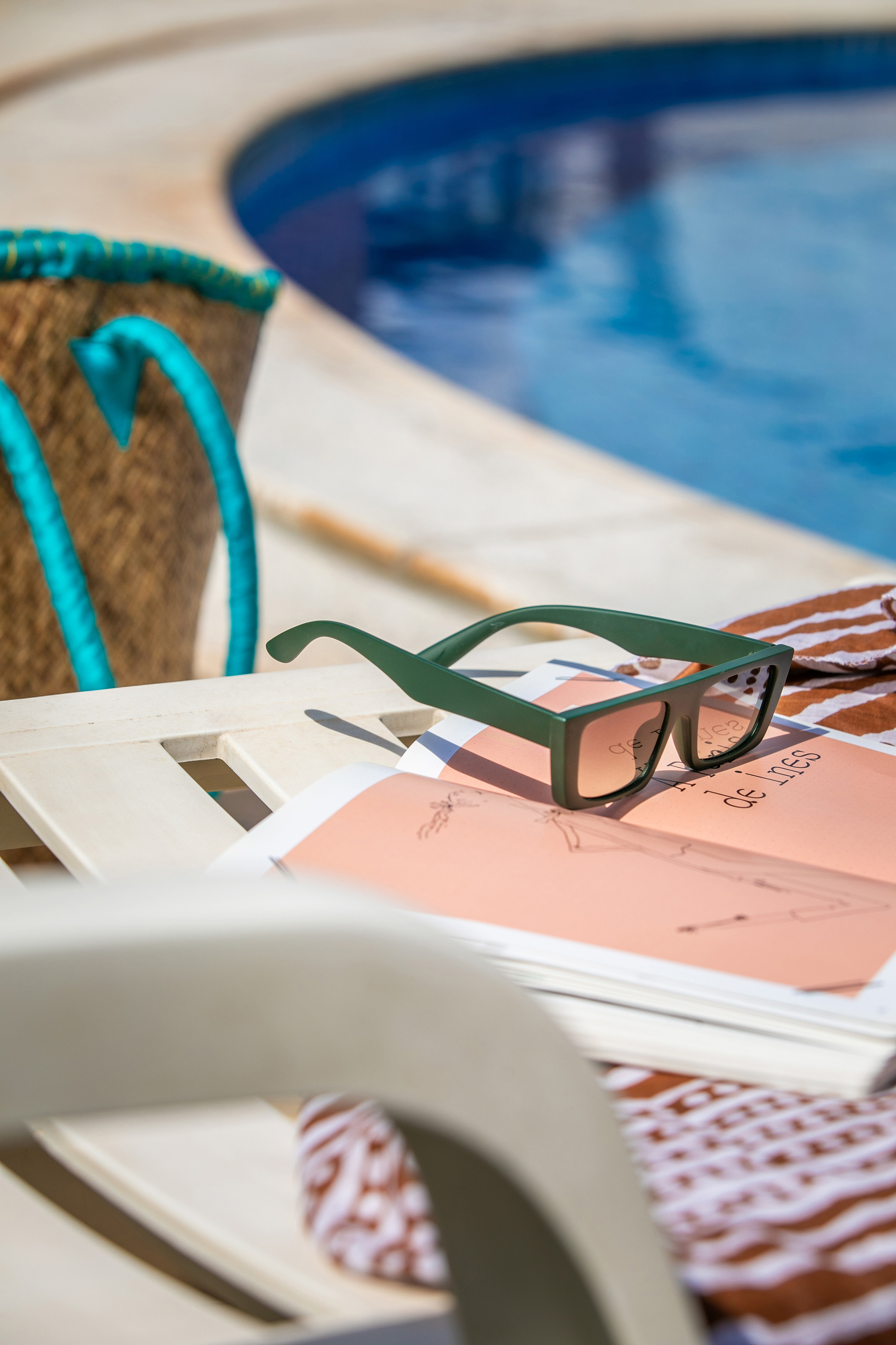 Green sunglasses and book on a sun lounger beside a clear blue pool.