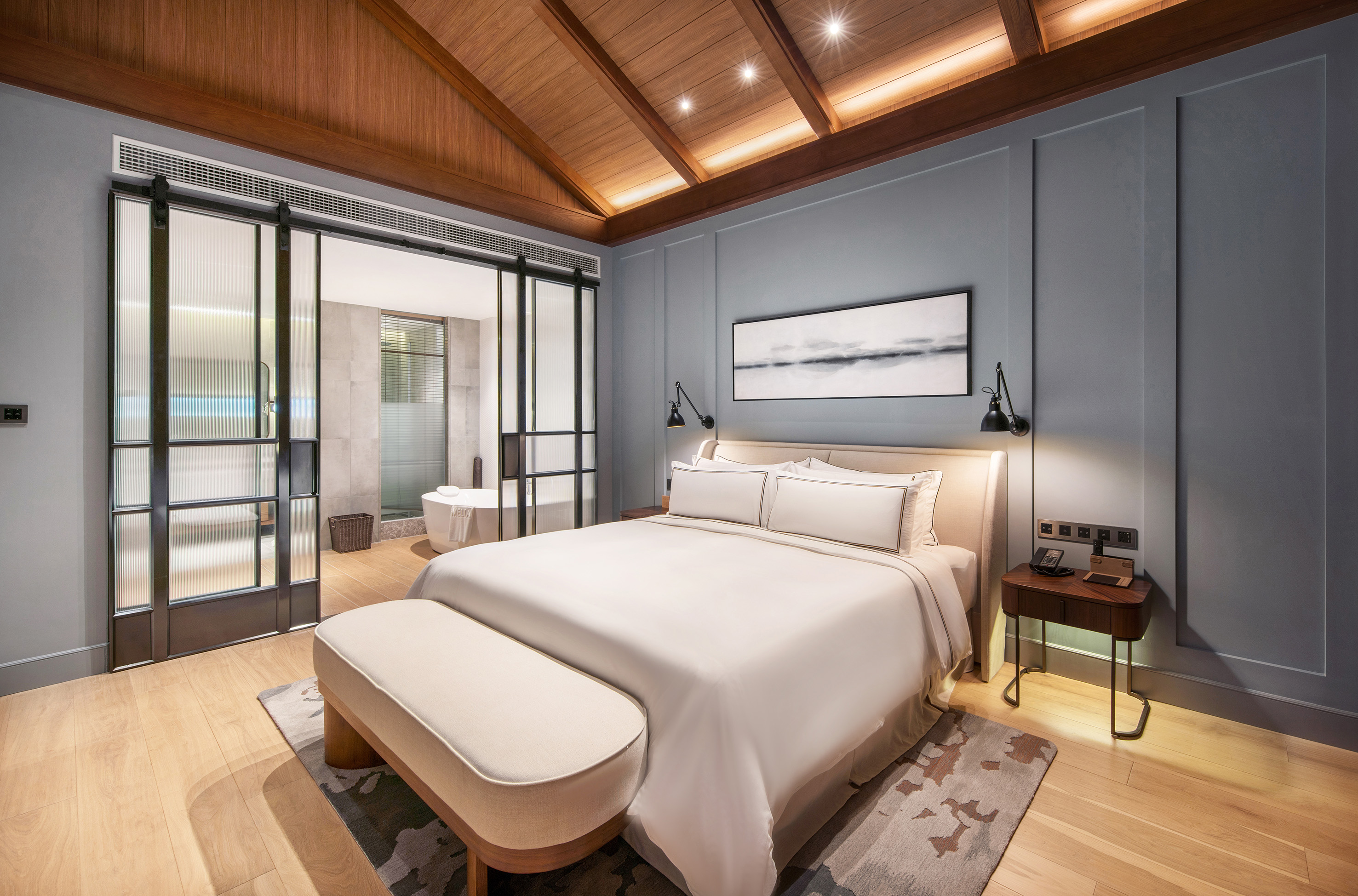 Modern hotel suite with wood ceiling, large bed, and glass sliding doors to bathroom.