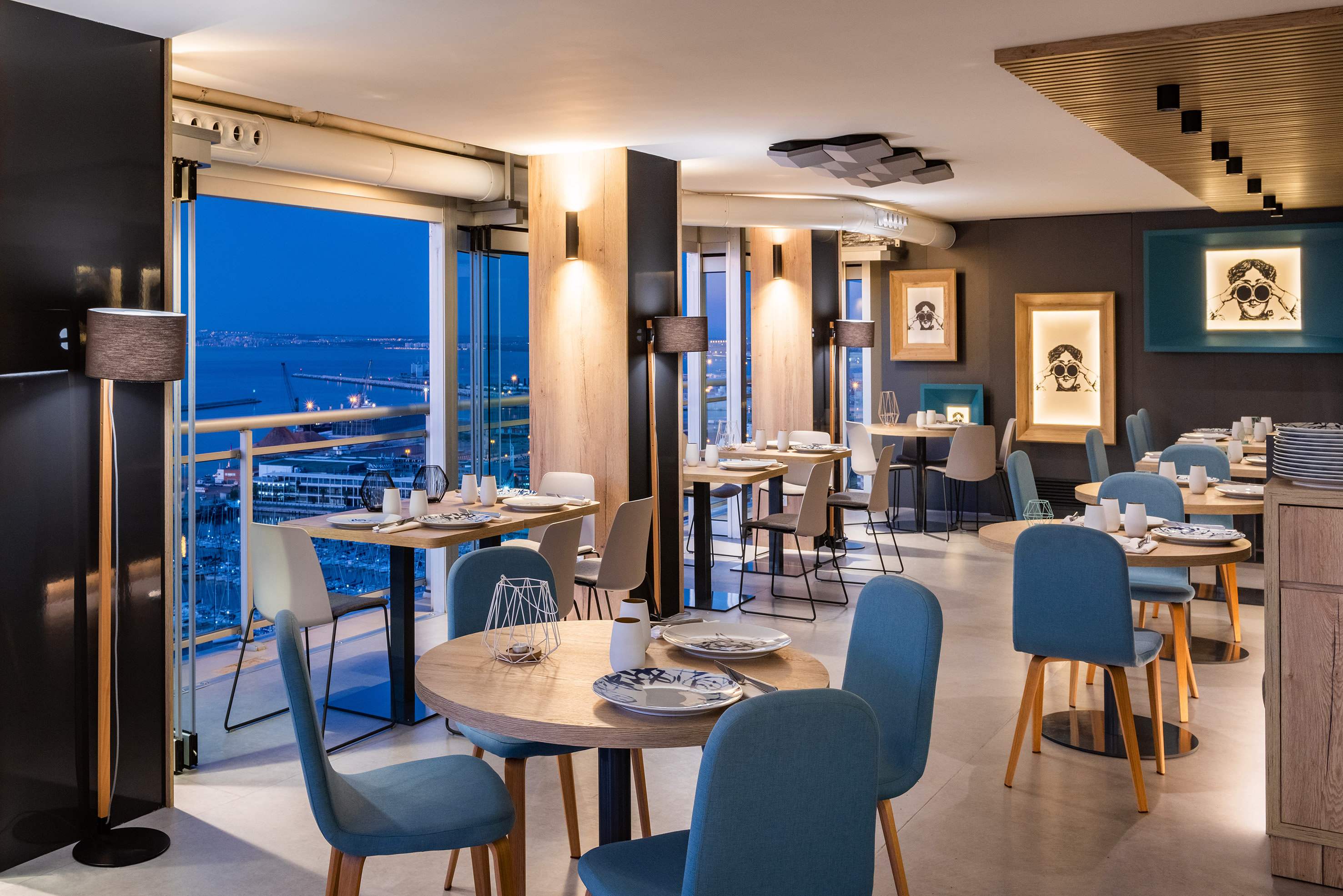 Modern restaurant dining room with blue chairs, wood decor, and night city ocean view.