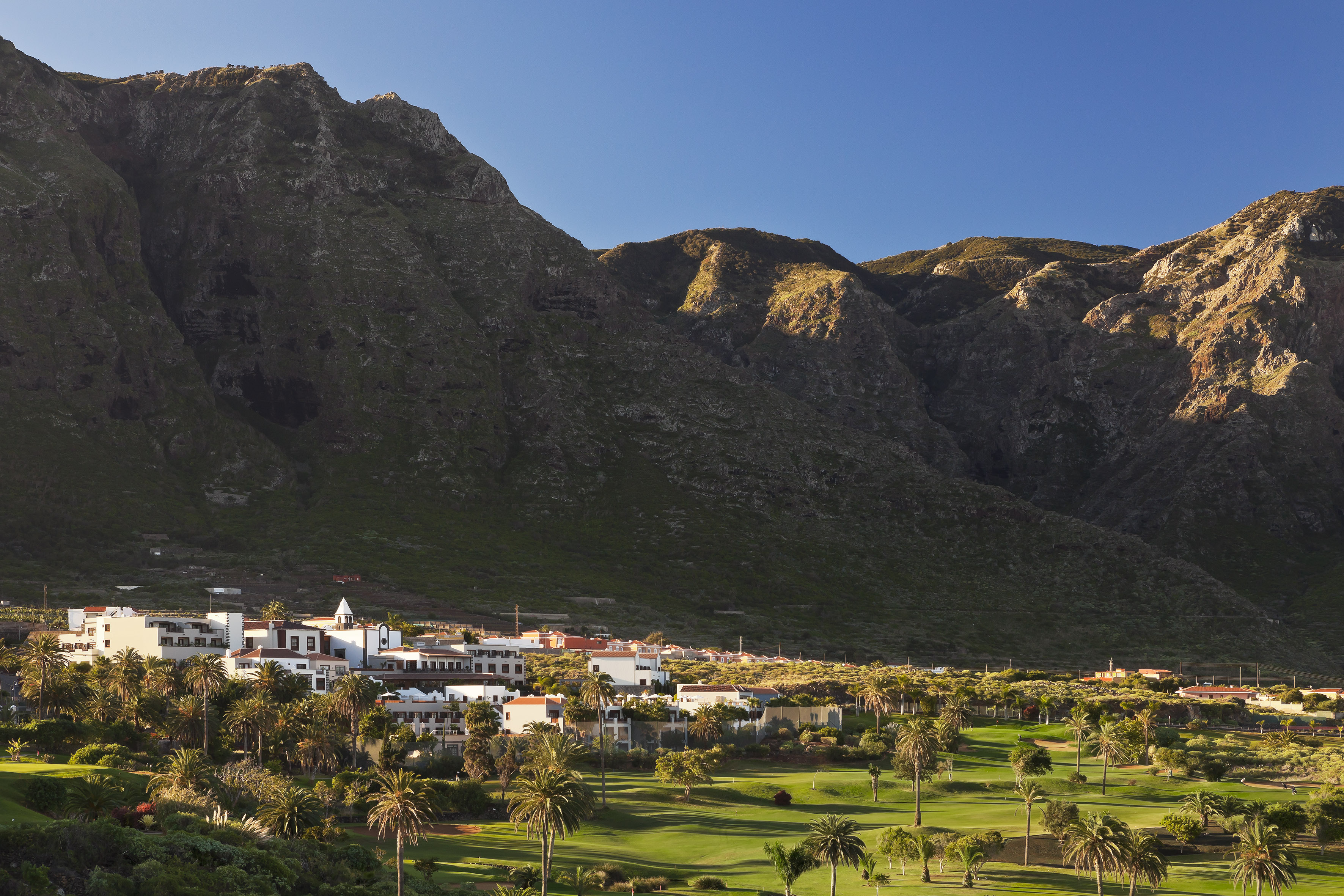 Lush golf course, palm trees, and resort buildings against rugged mountains.