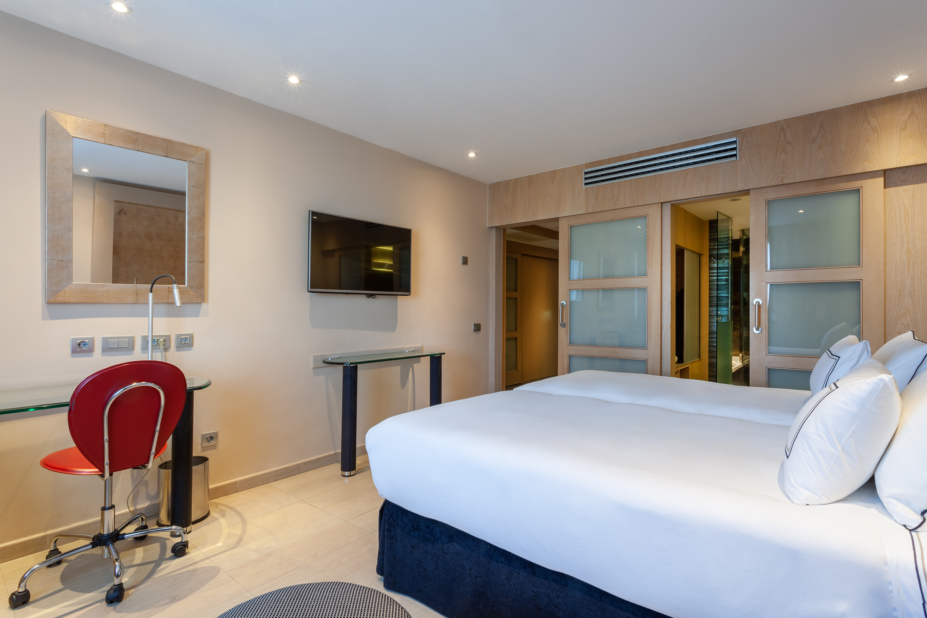 Modern hotel room with white beds, wall TV, desk, and sliding bathroom door.