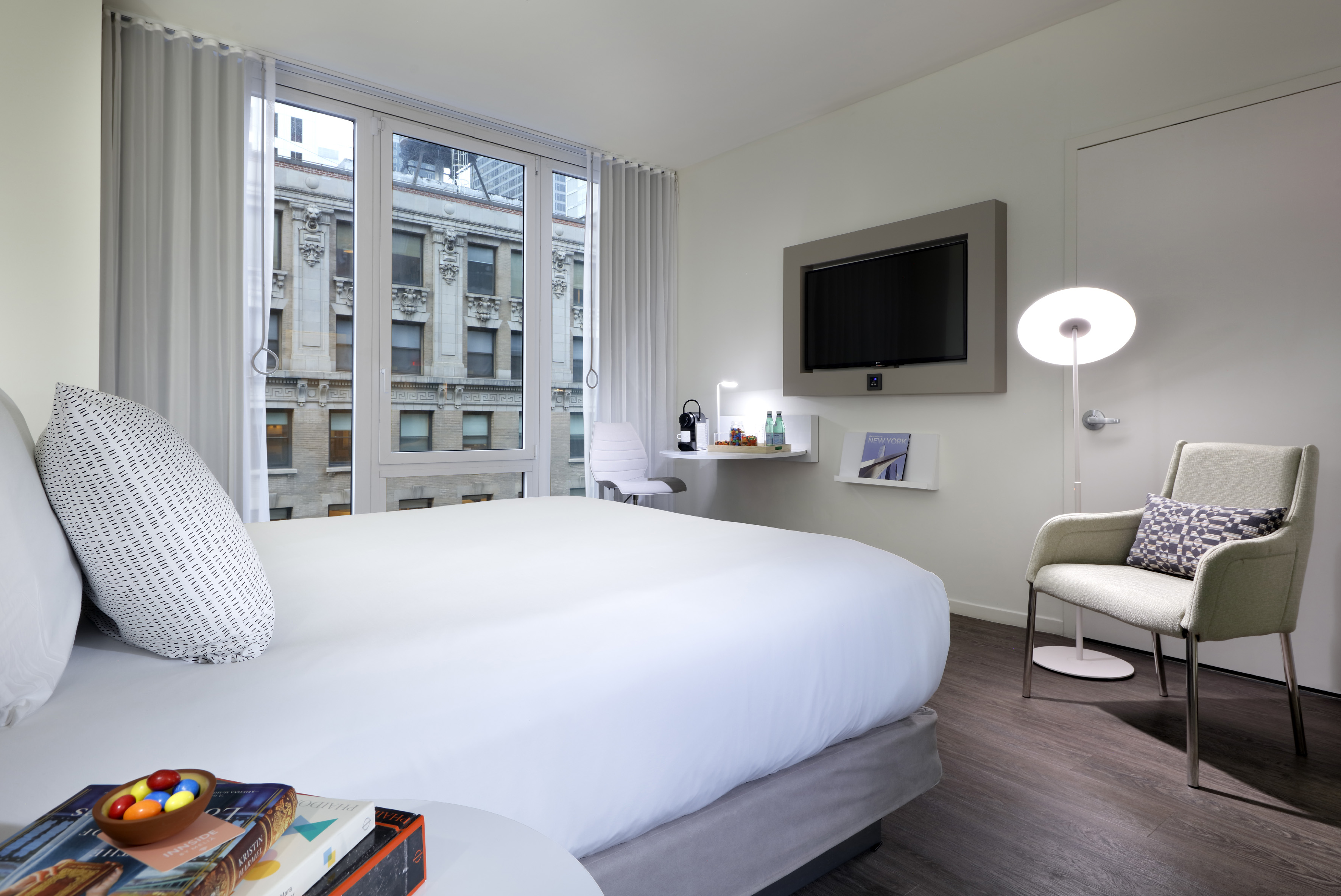 Modern hotel room with large bed, city view window, TV, and desk.