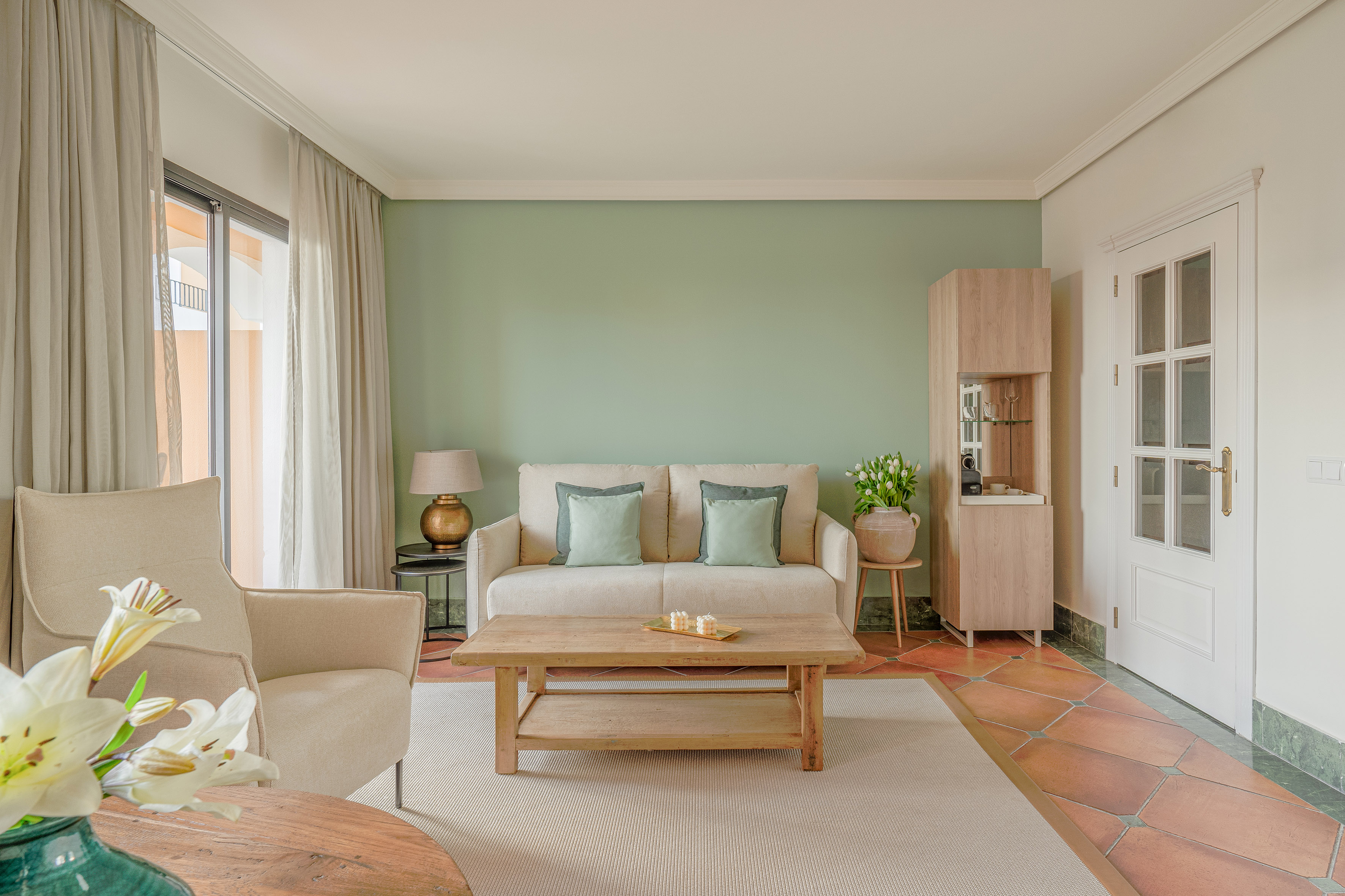 Cozy living room with green wall, beige sofa, wood coffee table, terracotta floor.