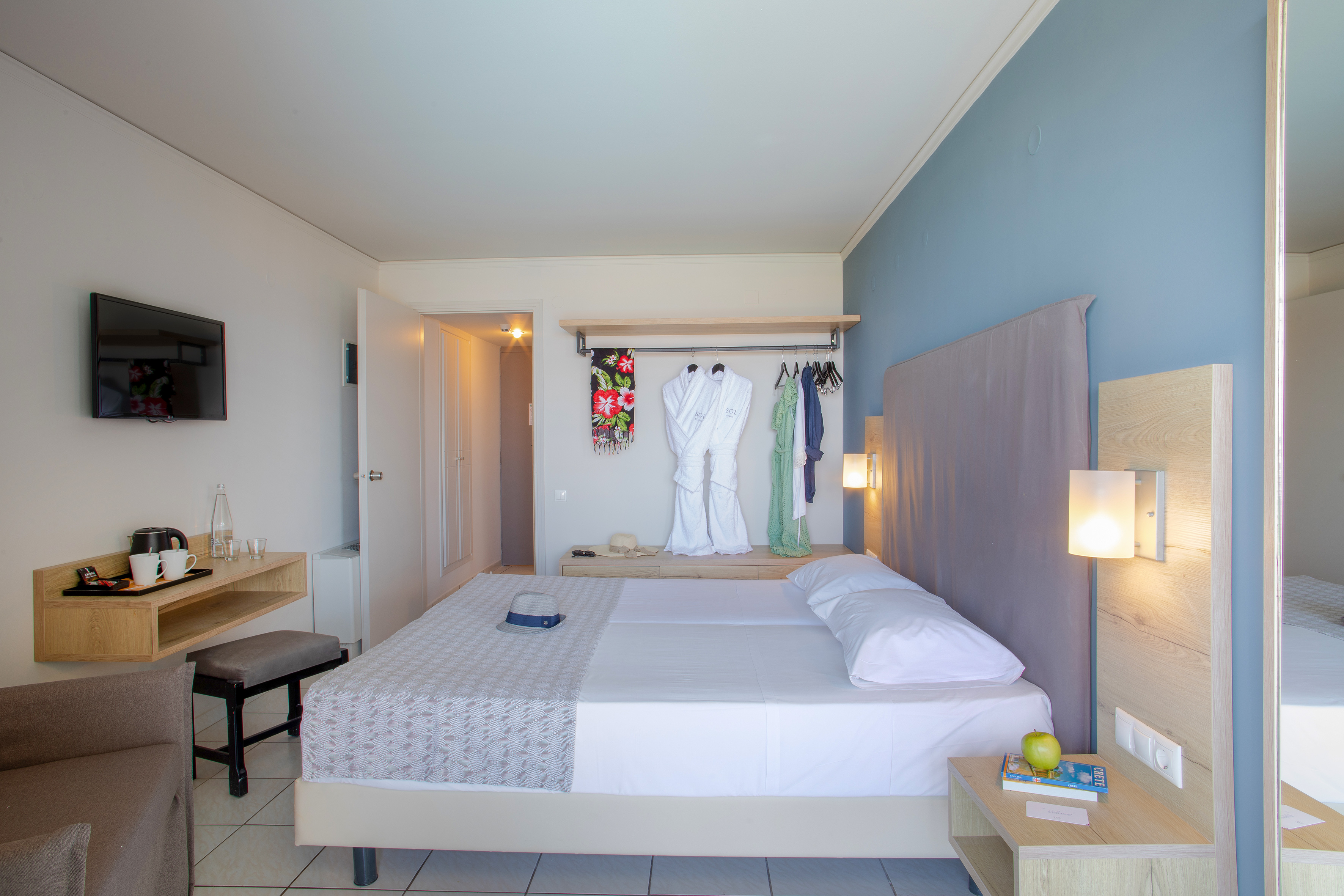 Modern hotel bedroom with twin beds, wall TV, and open wardrobe with robes.