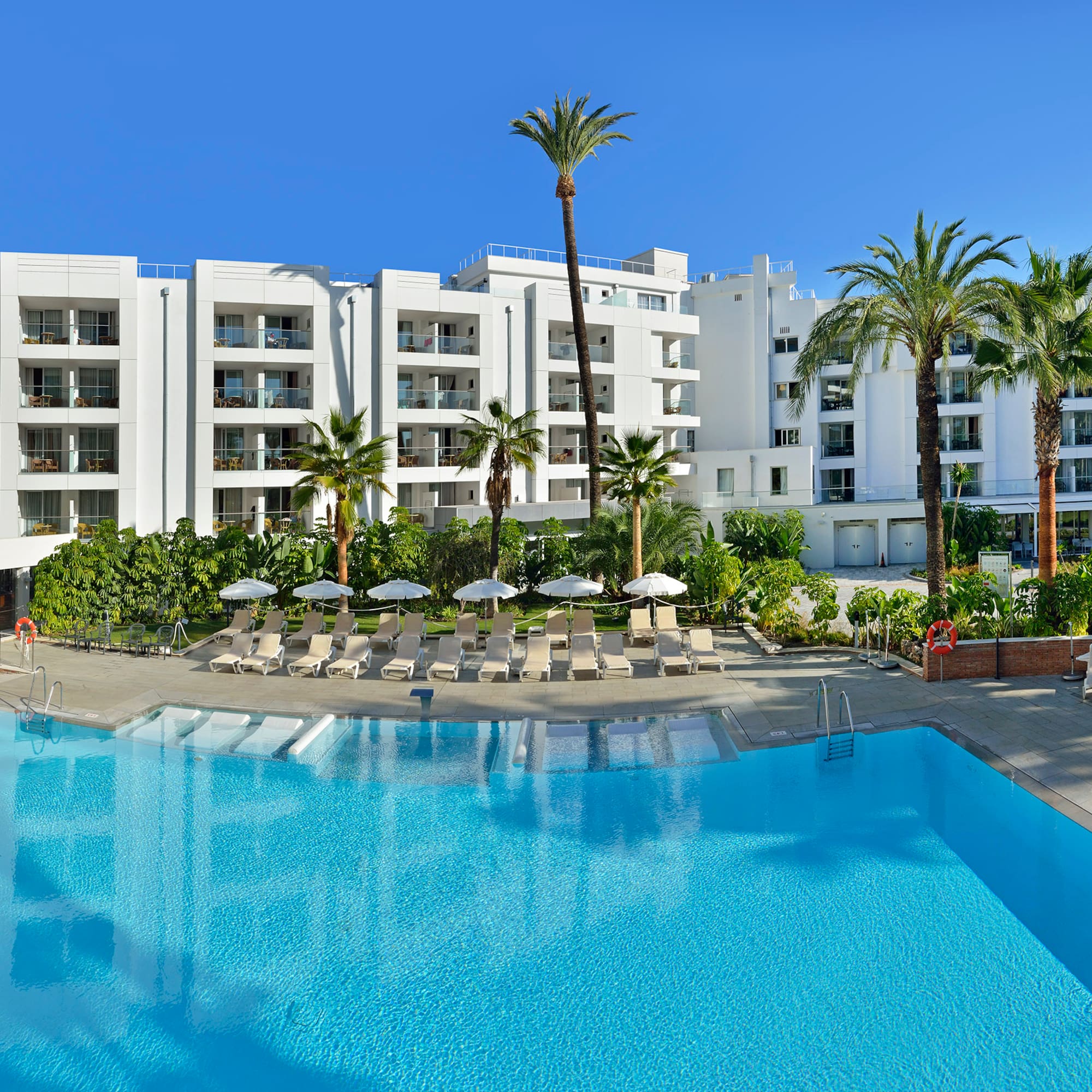 White resort hotel, large swimming pool, palm trees, and sun loungers.